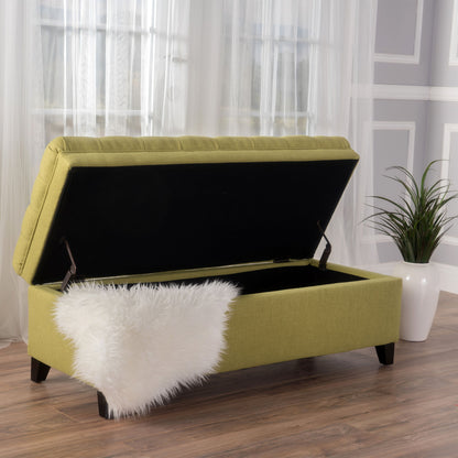 The Juliana Storage Ottoman with birch legs is shown in green upholstery, its lid open to reveal a black interior. A white faux fur throw drapes over the side, and the ottoman sits on wooden floors near a window with a nearby potted plant.