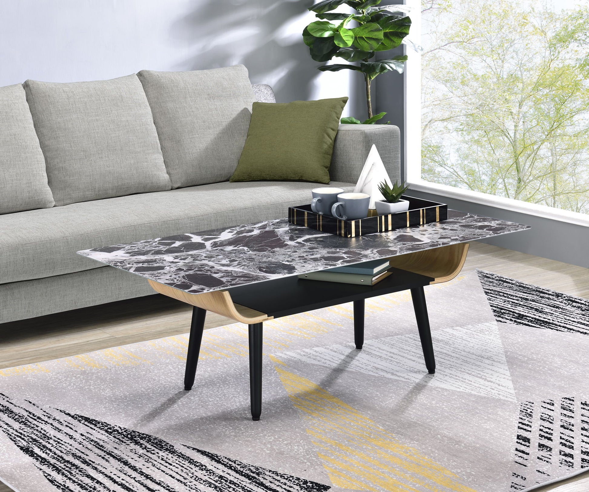 A modern living room with a gray sofa, green pillow, Landon Coffee Table featuring a glass marble texture top and bent wood design holding mugs and decor, patterned rug, potted plant, and large window overlooking leafy trees.