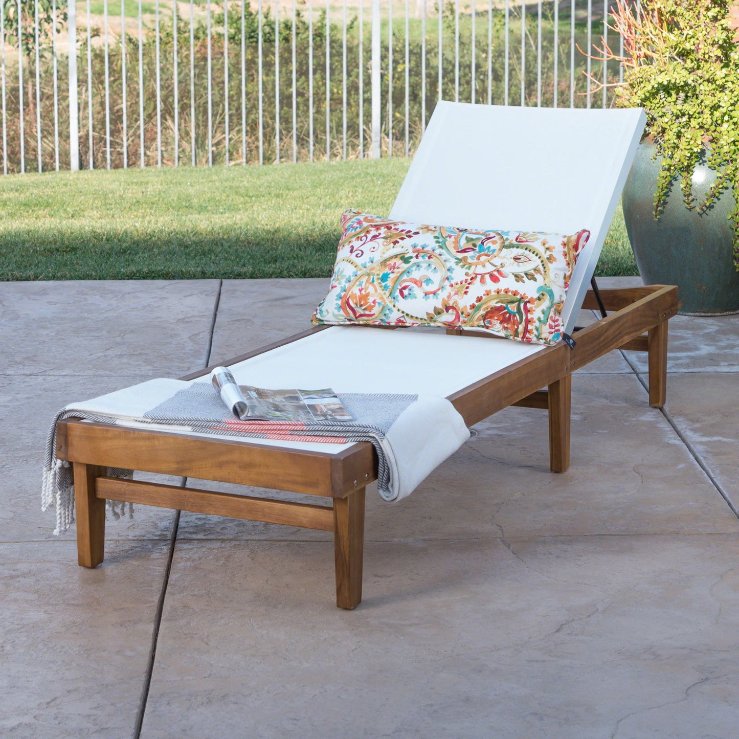 An Outdoor Chaise Lounge With Acacia Wood Frame - White / Teak sits on a stone patio near a grassy lawn, accented with a white cushion, colorful pillow, gray-striped throw, and set against a backdrop of greenery and a metal fence.