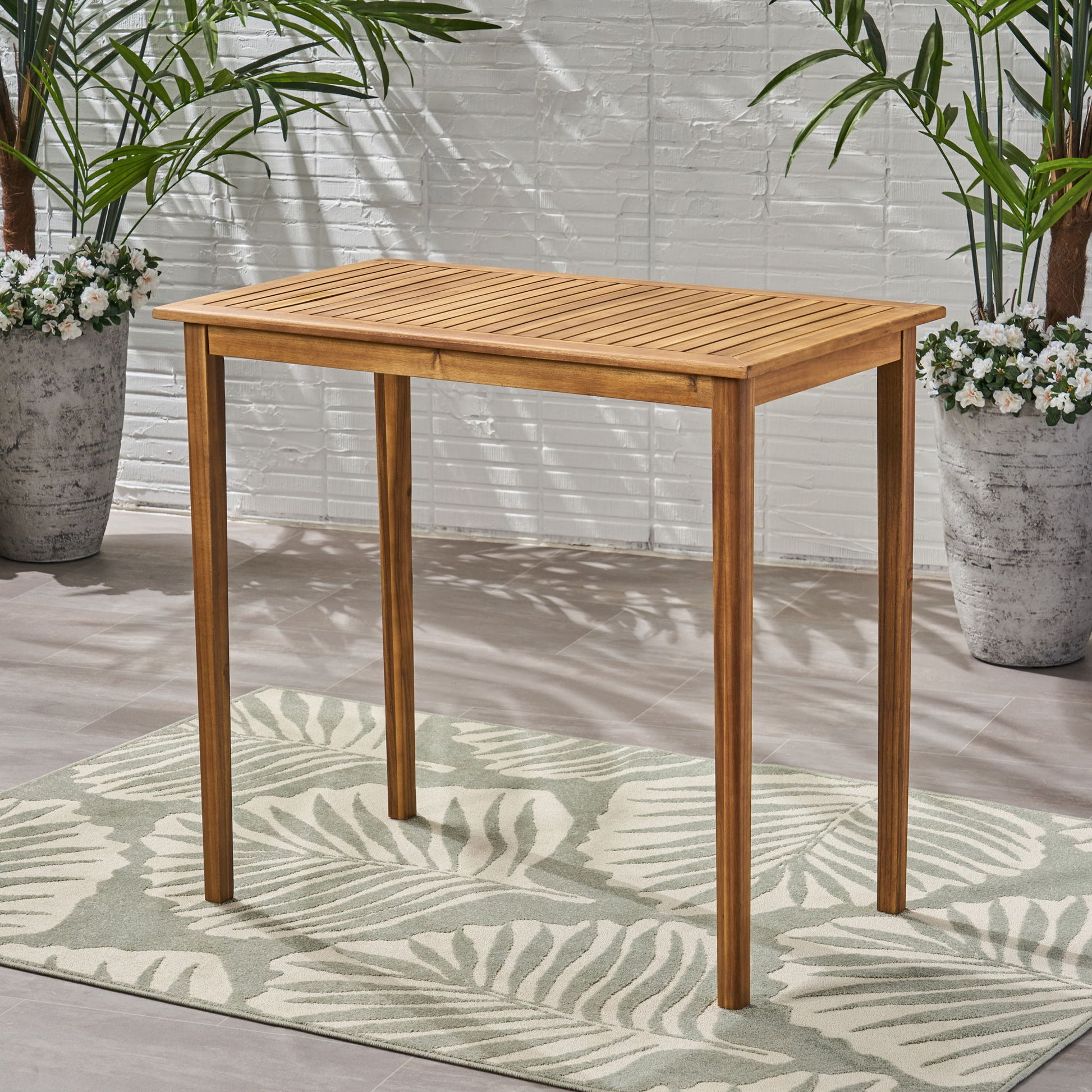 The Outdoor Minimalist Acacia Wood Rectangle Bar Table with a slatted top stands on a light green leaf-patterned rug, surrounded by potted plants and set against a white brick wall.
