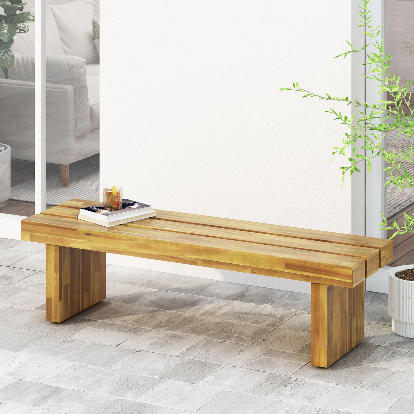 The Nahanni Rustic Acacia Wood Bench with Slat Design in Natural sits on a stone patio next to a potted plant, topped with books and a drink. Sliding glass doors open to reveal a cozy living room with a sofa in the background.