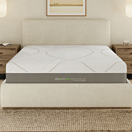 A neatly made bed features the 12" Profile Massage Mattress Hybrid on a cream-colored upholstered frame, flanked by wooden nightstands and lit by wall sconces. A framed picture hangs above the headboard.