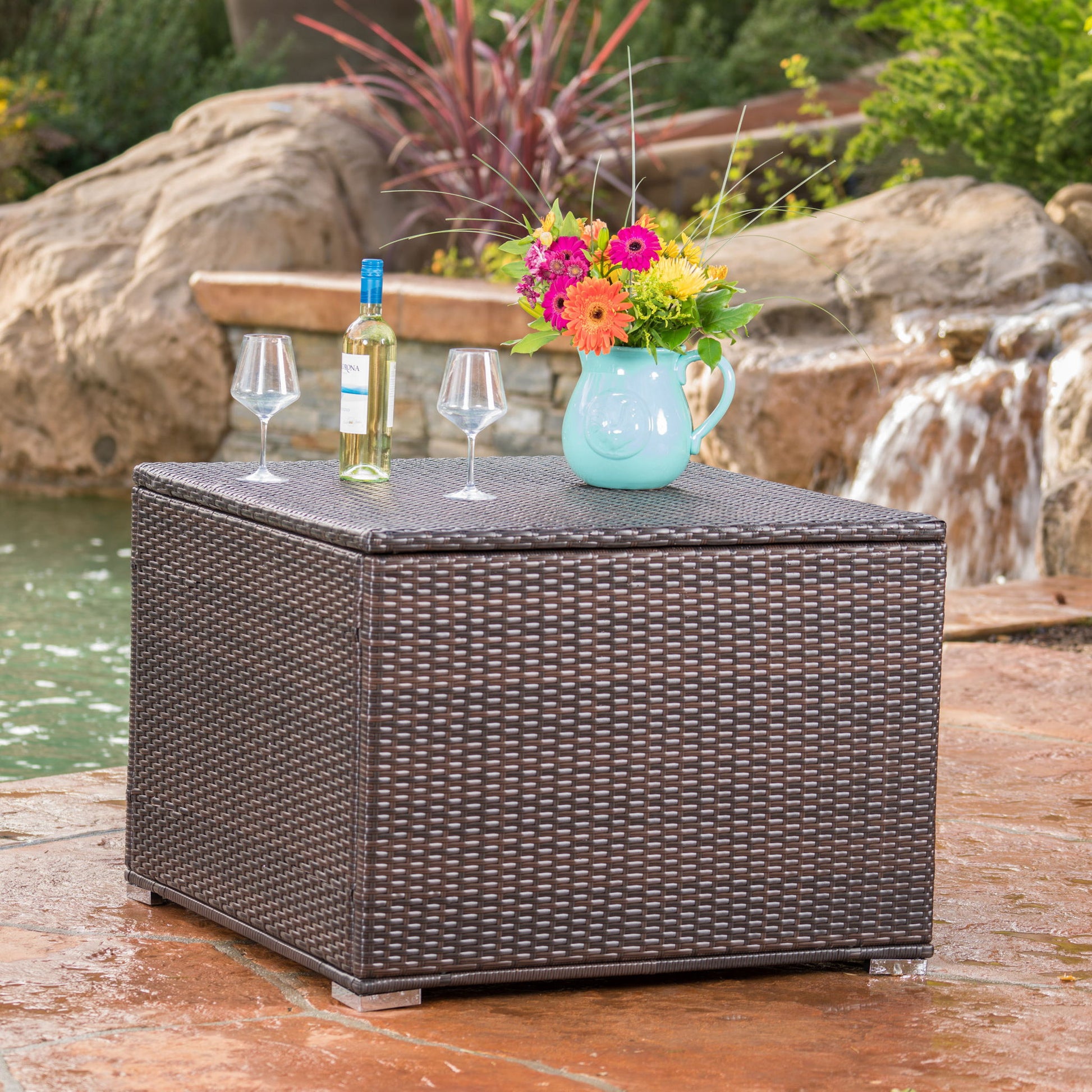 The Santa Rosa - Storage Table is styled with a bottle of wine, two glasses, and a turquoise pitcher of flowers, set next to a tranquil garden waterfall and pond.