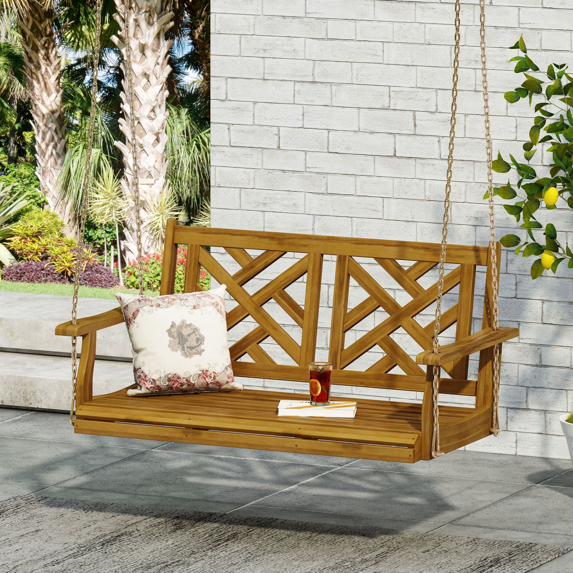 The Alejandro Acacia Wood Porch Swing with slat panel seating hangs from chains. On its seat are a floral pillow, a book, and iced tea. The swing sits on a patio with a brick wall, lush greenery, and palm trees in the background.