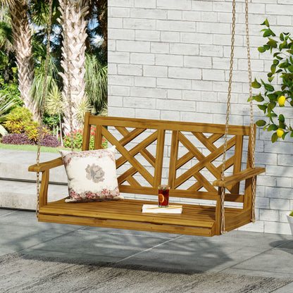 The Alejandro Acacia Wood Porch Swing with slat panel seating hangs from chains. On its seat are a floral pillow, a book, and iced tea. The swing sits on a patio with a brick wall, lush greenery, and palm trees in the background.
