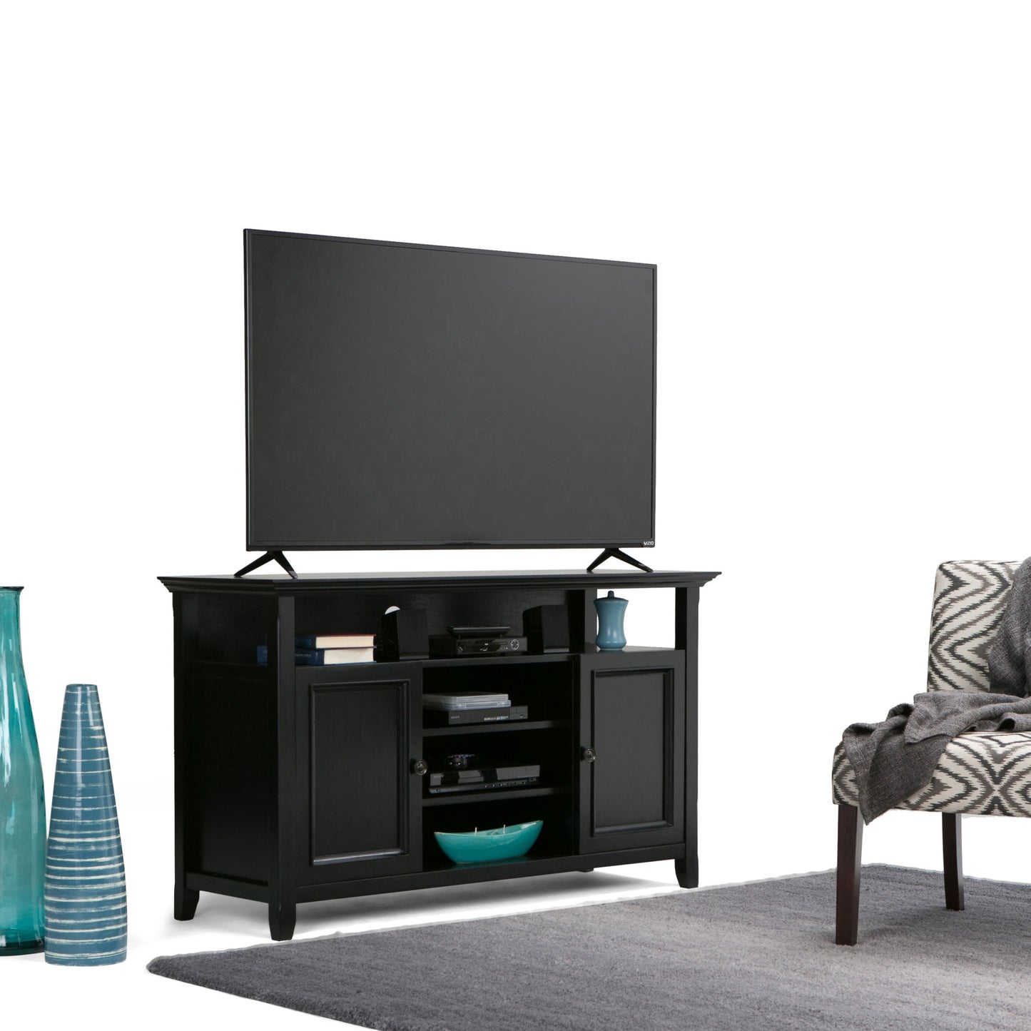 The Amherst Handcrafted TV Media Stand, finished in black, supports a flat-screen TV and displays decor and electronics on its shelves and cabinets. Blue vases, a patterned chair, and a gray rug complete the room’s look.