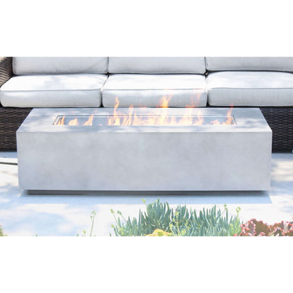 A Modern Concrete Design Propane Outdoor Fire Pit Table with visible flames is placed in front of a light gray outdoor sofa on the patio, surrounded by lush greenery and blooming flowers.