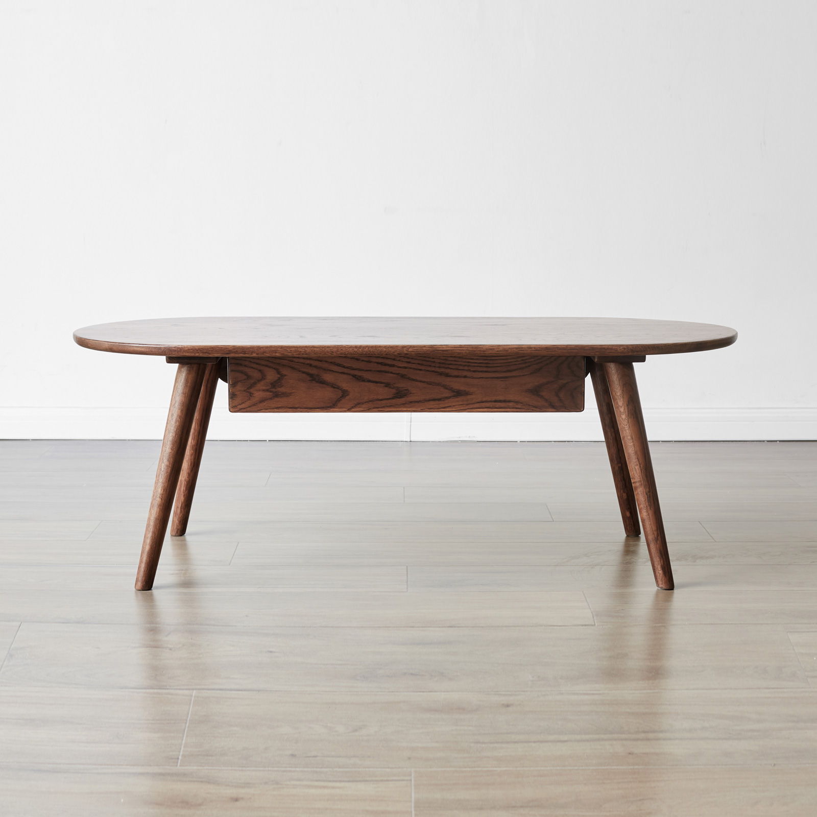 The Capsule Centre Low Table With Drawers Coffee Table features a low, oval wooden design with splayed legs, perfect for placing against a plain white wall and light wood floor.