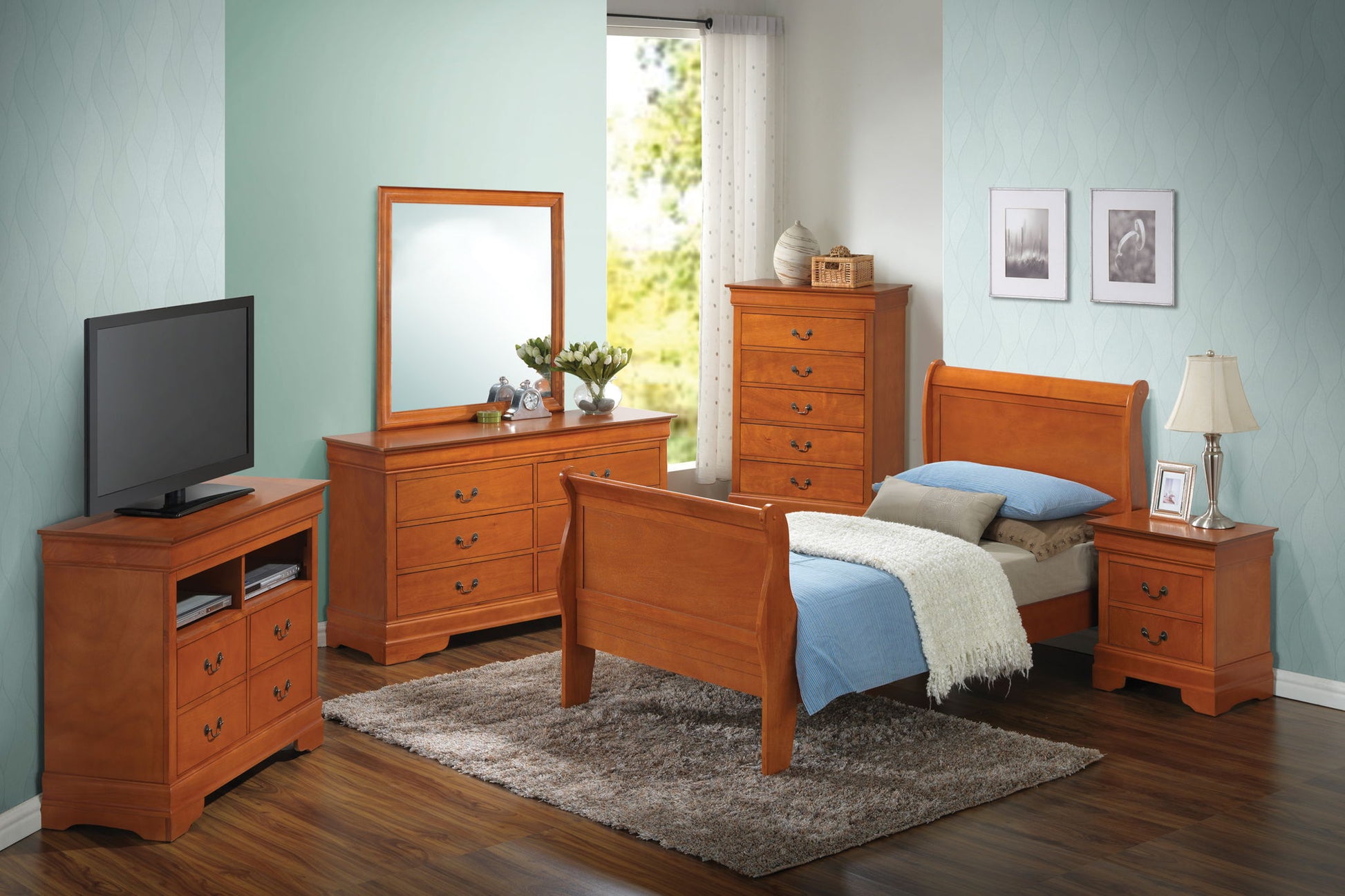 A bedroom with light blue walls features a Panel Sleigh Bed with blue bedding, wood furniture including a dresser with mirror, tall chest, nightstand, and TV stand. A lamp and framed pictures decorate the space.
