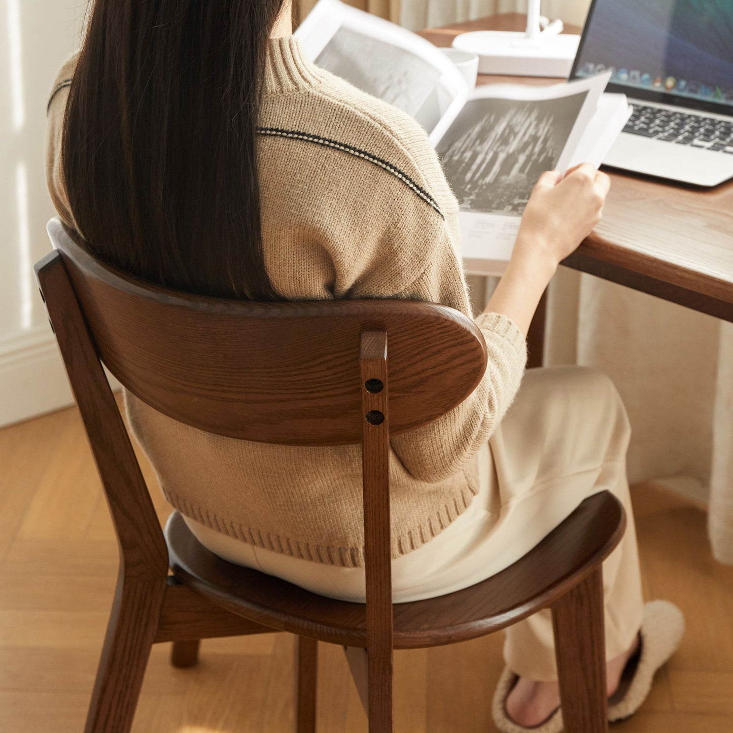 A person with long dark hair, wearing a beige sweater and light pants, sits on a Simple Dining Chair at a table reading a book, with an open laptop and another book nearby.