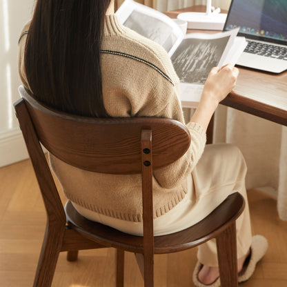 A person with long dark hair, wearing a beige sweater and light pants, sits on a Simple Dining Chair at a table reading a book, with an open laptop and another book nearby.