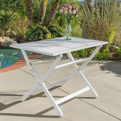 The Blissique Outdoor Foldable Wood Dining Table with Slat Design, in white, sits on a patio near the pool topped with a glass vase of pink flowers, surrounded by lush green plants and trees.