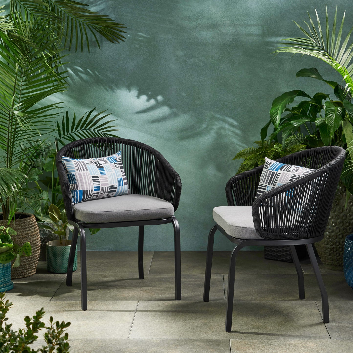 Two Outdoor Wicker Chairs with powder-coated iron legs, gray cushions, and patterned pillows sit on a tiled patio, surrounded by green potted plants against a textured green wall.