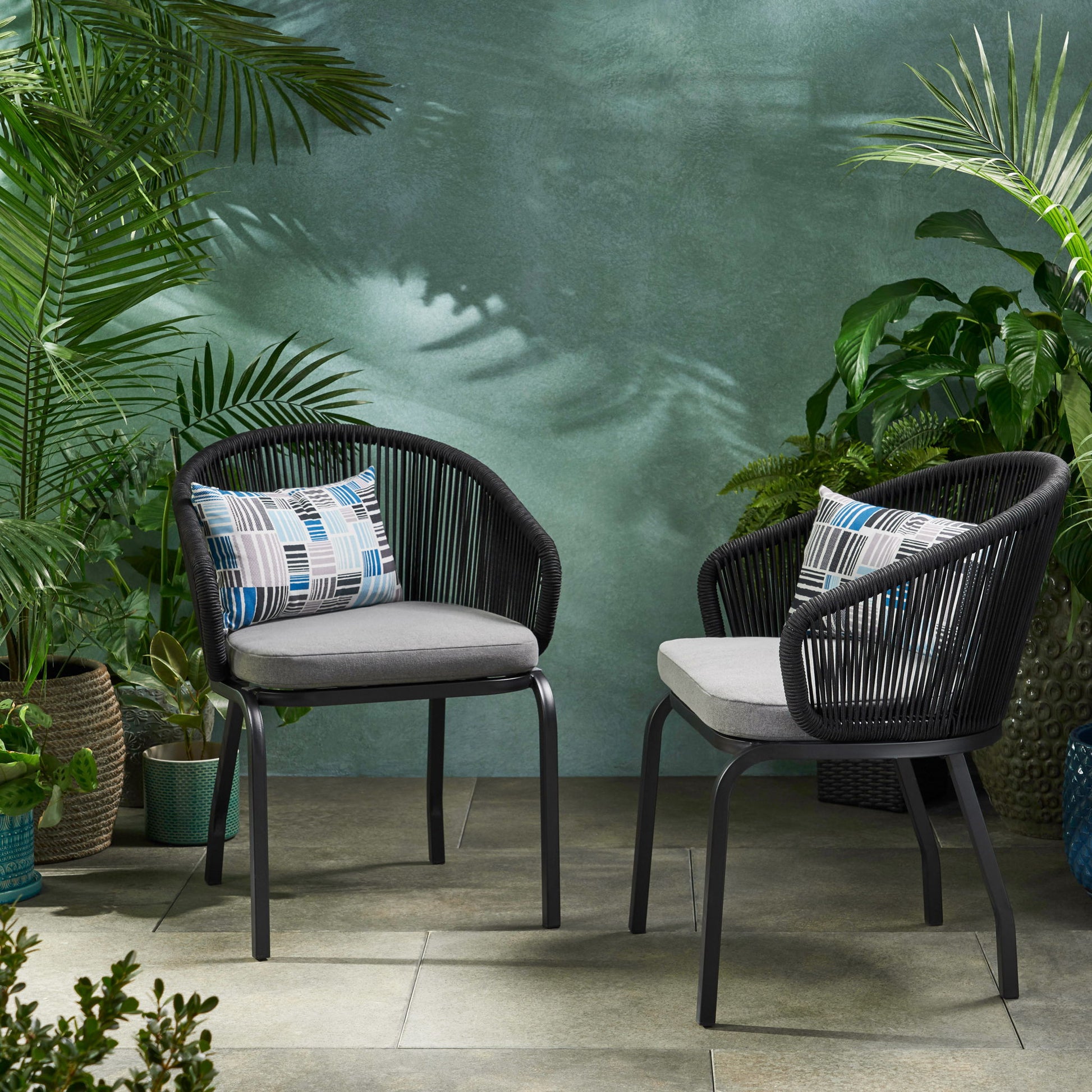 Two Outdoor Wicker Chairs with powder-coated iron legs, gray cushions, and patterned pillows sit on a tiled patio, surrounded by green potted plants against a textured green wall.