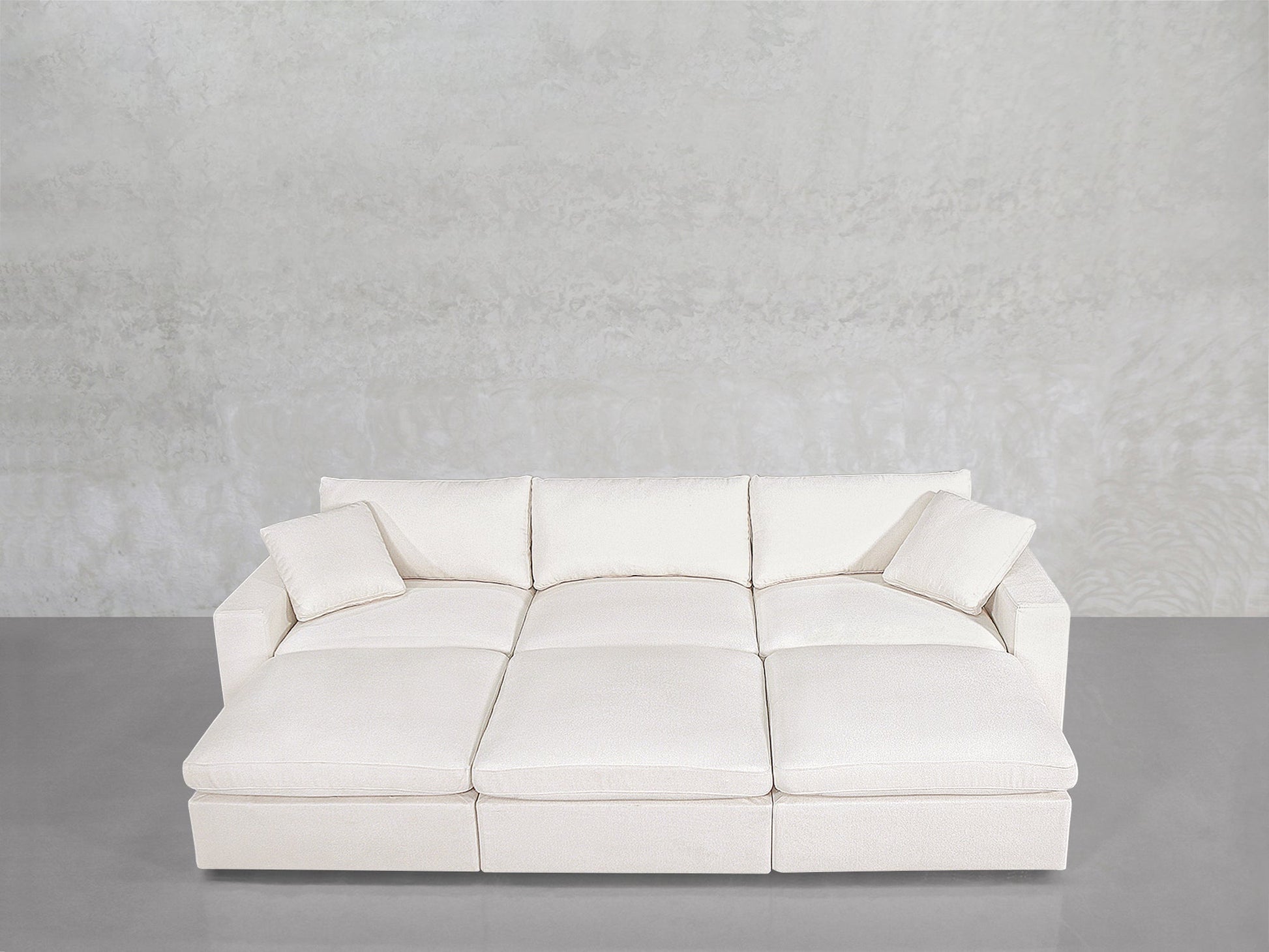 A modern, minimalist 6-Seat Modular Daybed in white, featuring three seat cushions, three back cushions, and two matching throw pillows, sits against a light gray textured wall on a smooth gray floor.
