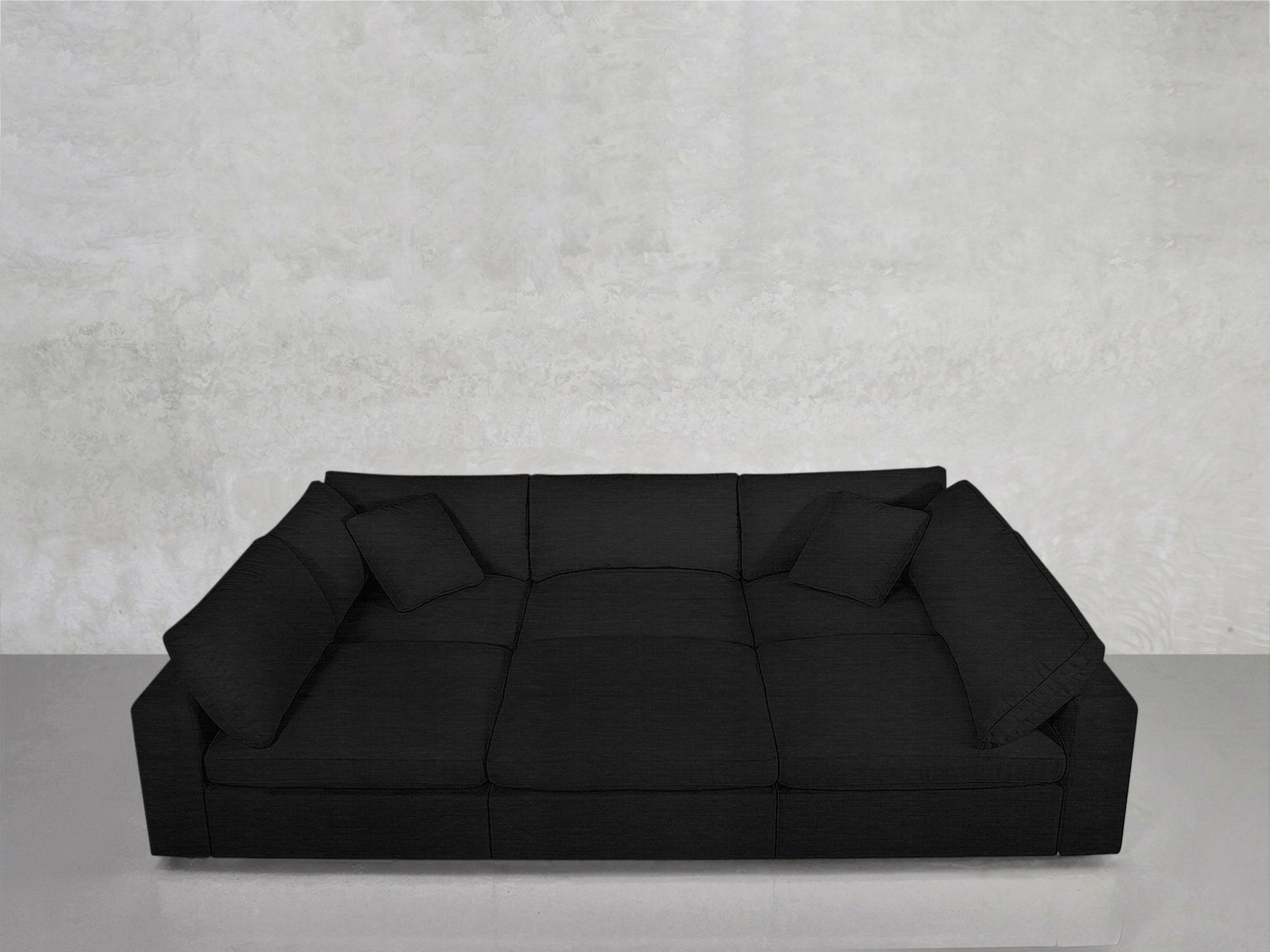 A 6-Seat Modular Pit in black with wide armrests and two matching cushions, set against a light grey textured wall on a smooth, light-colored floor.