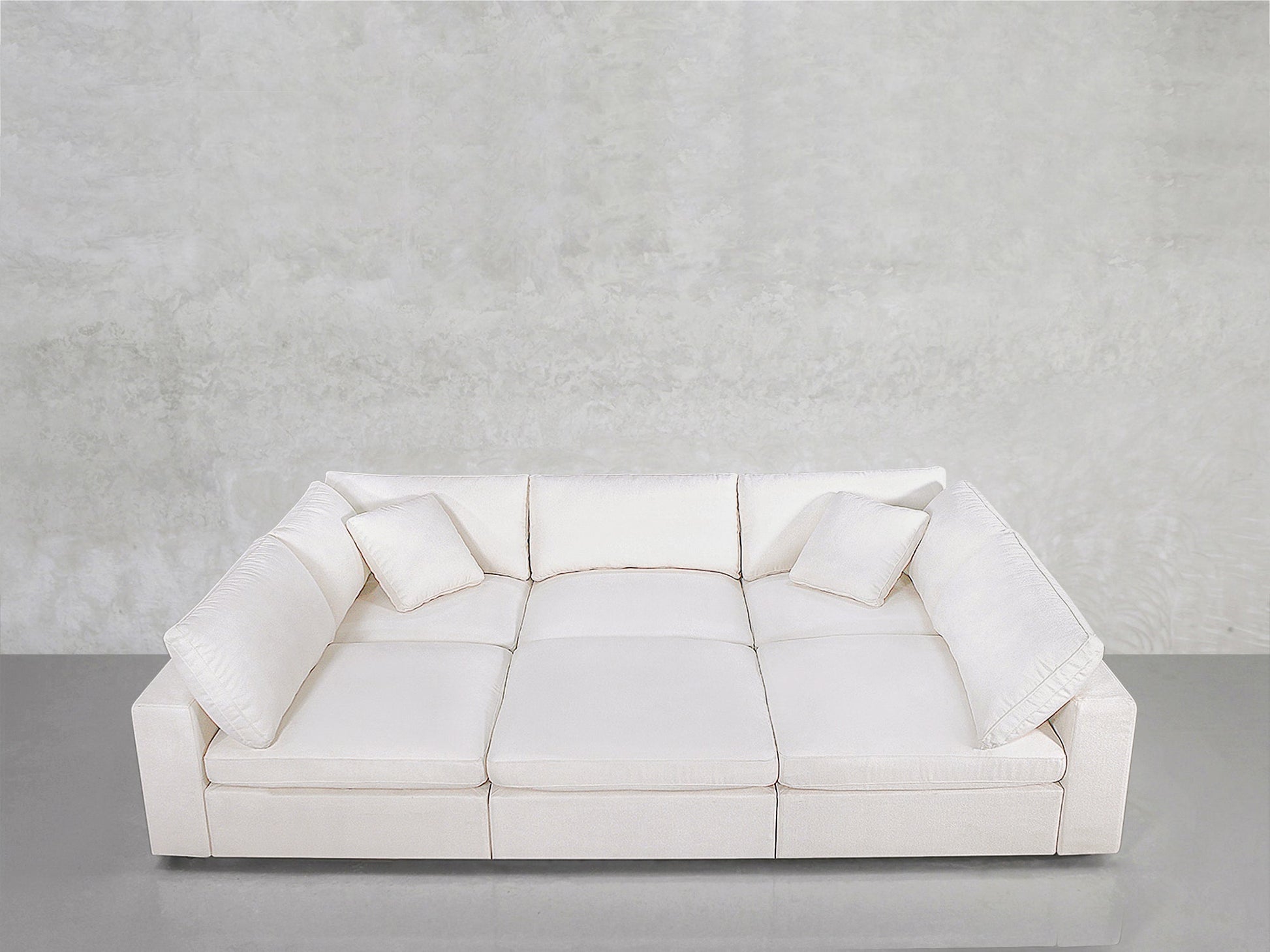 A contemporary white 6-Seat Modular Pit with three seat cushions and four matching throw pillows, positioned near a light gray textured wall and reflective gray floor.