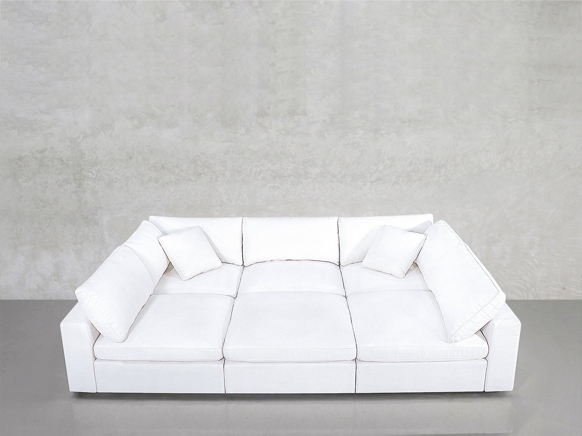 A modern white 6-Seat Modular Pit with three seat cushions and four throw pillows sits against a plain, light gray textured wall on a smooth gray floor.