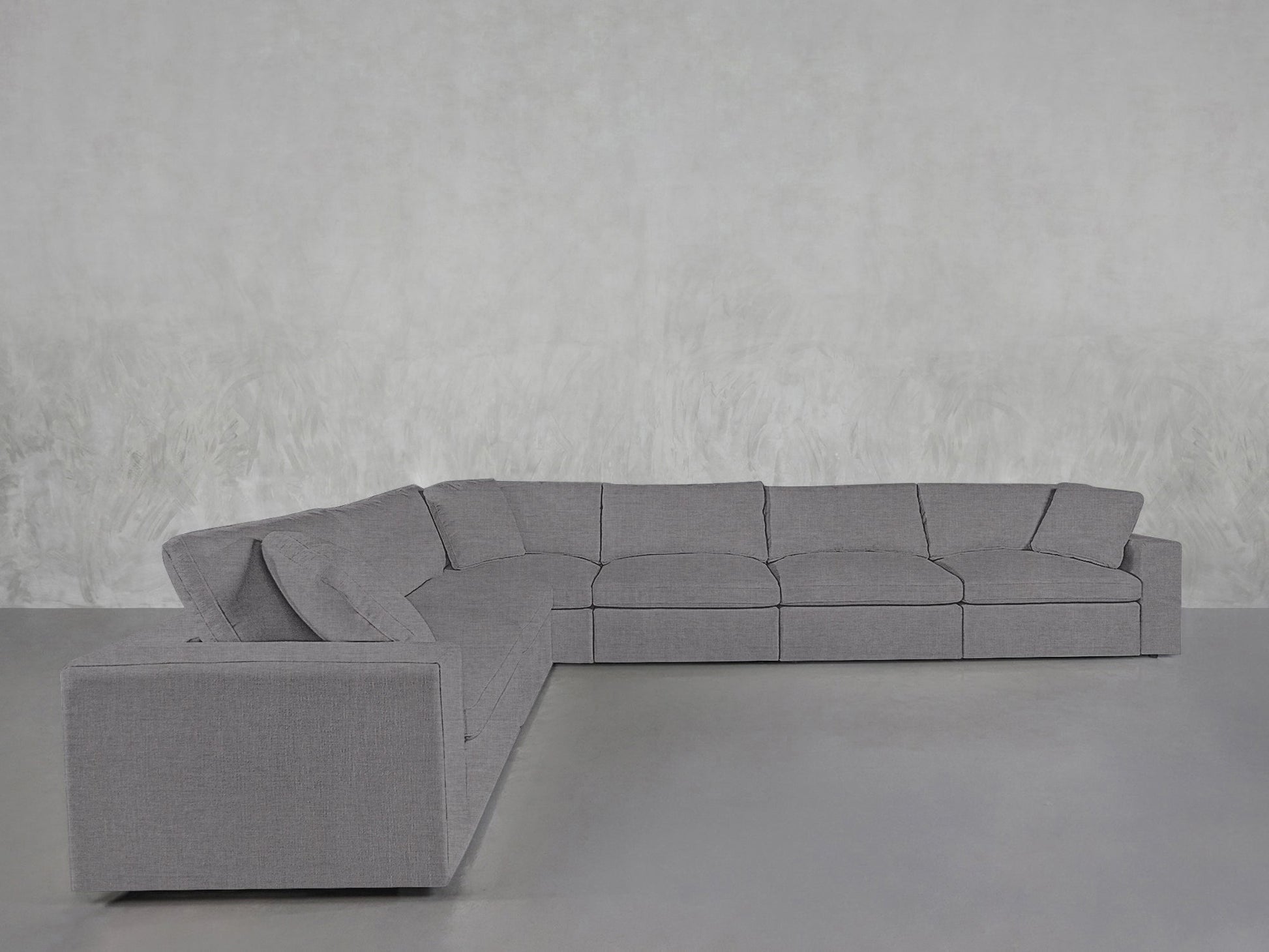 A 6-Seat Modular Corner Sectional with thick cushions and clean lines sits on a smooth gray floor against a light, textured wall, featuring a modern gray design and a left-facing chaise.