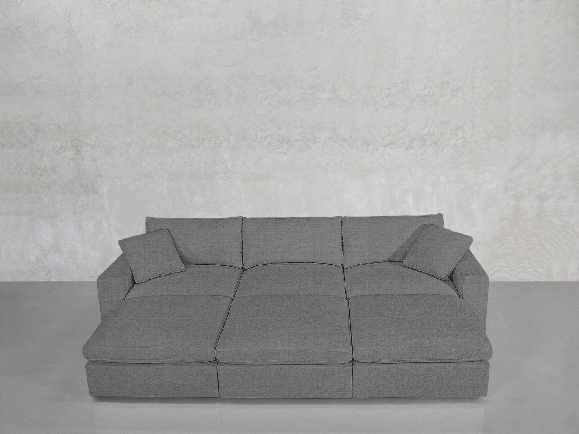 A contemporary 6-Seat Modular Daybed in gray, featuring three large seat cushions and four matching square back cushions, set against a plain light-textured wall on a smooth floor.