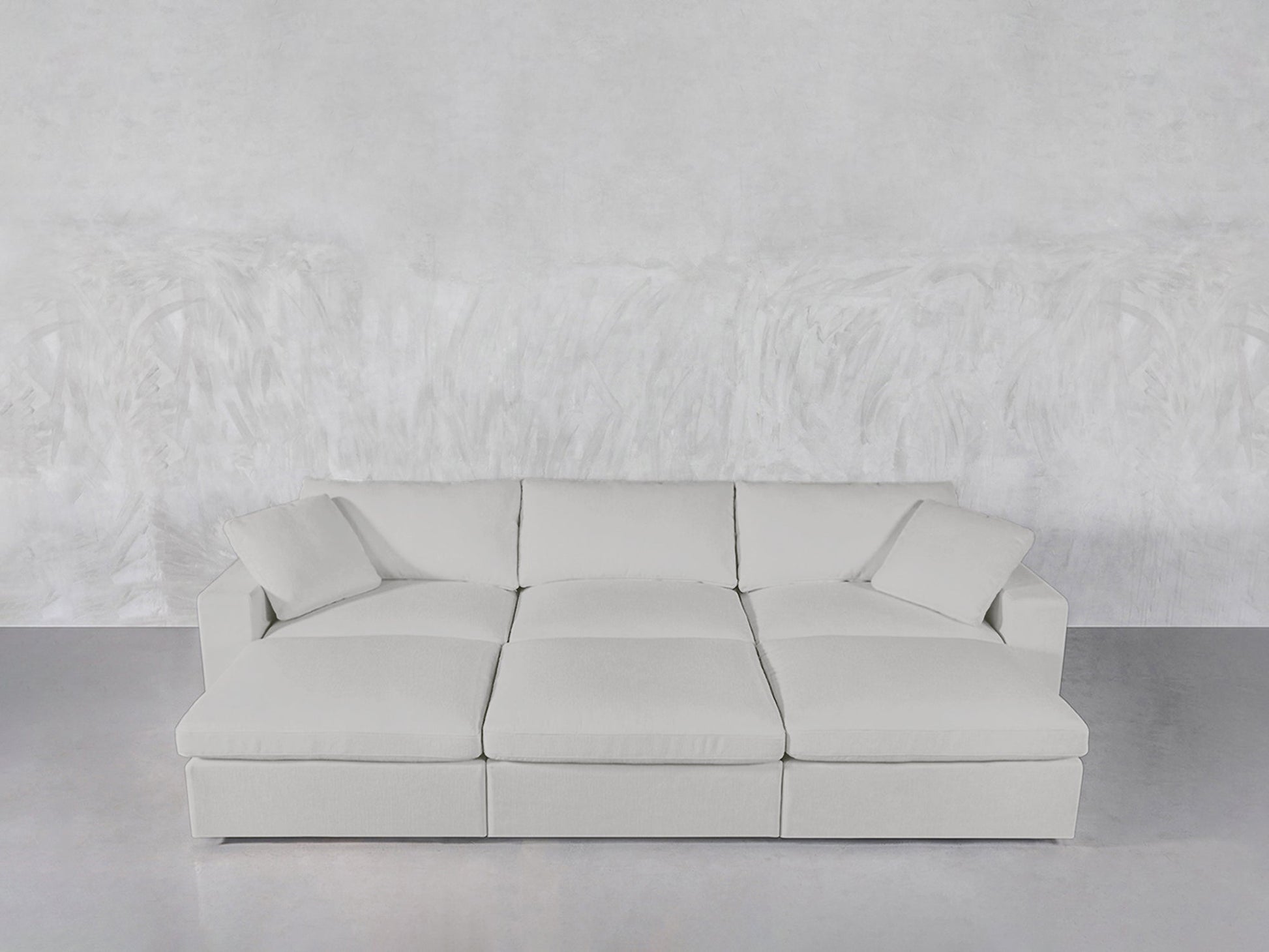 The 6-Seat Modular Daybed, in light gray with three large seat cushions and two pillows, is arranged against a textured white wall and smooth gray floor.