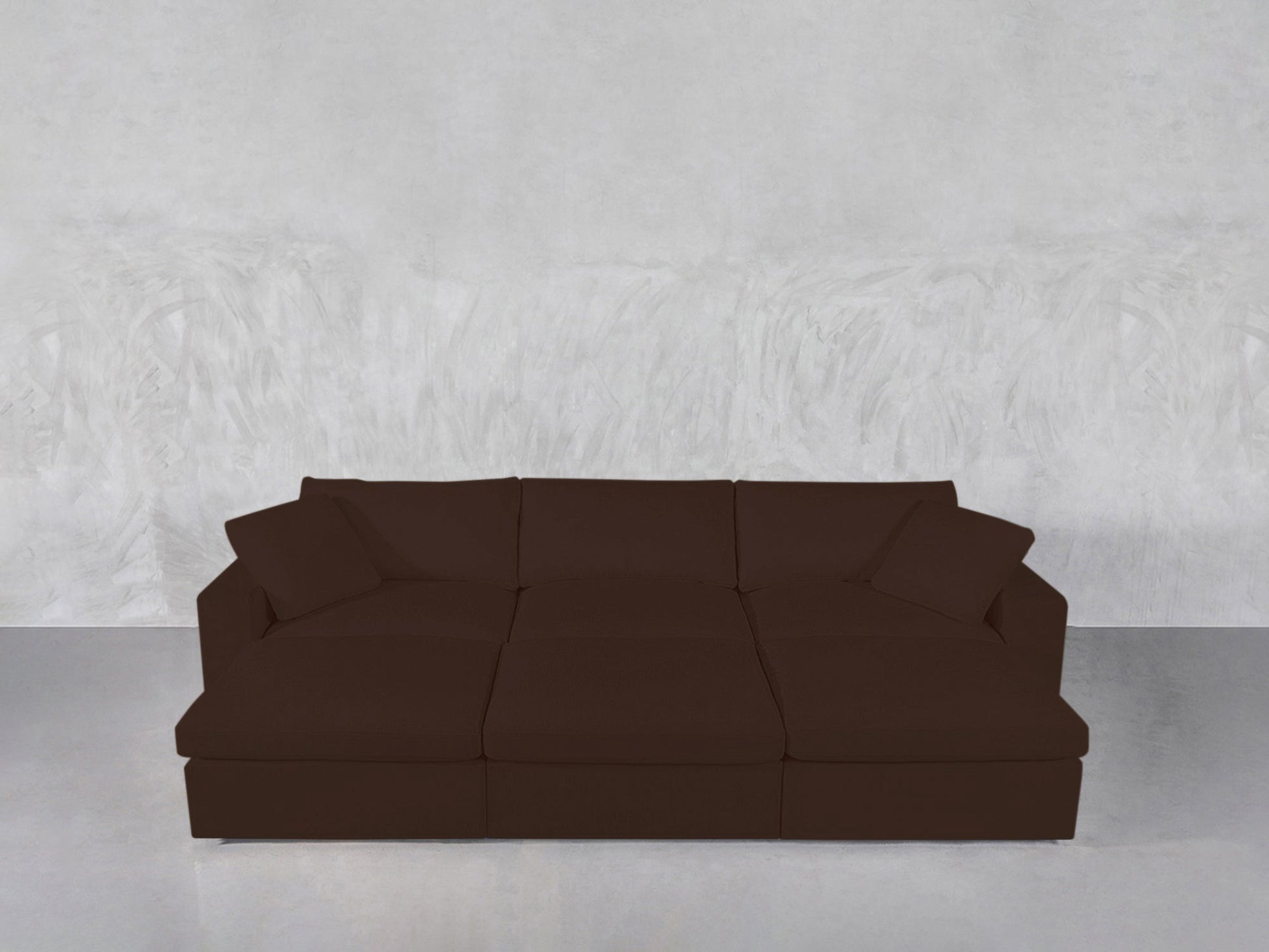 A 6-Seat Modular Daybed in dark brown, featuring three seat cushions and three back cushions, set against a plain light gray textured wall and a light gray floor.