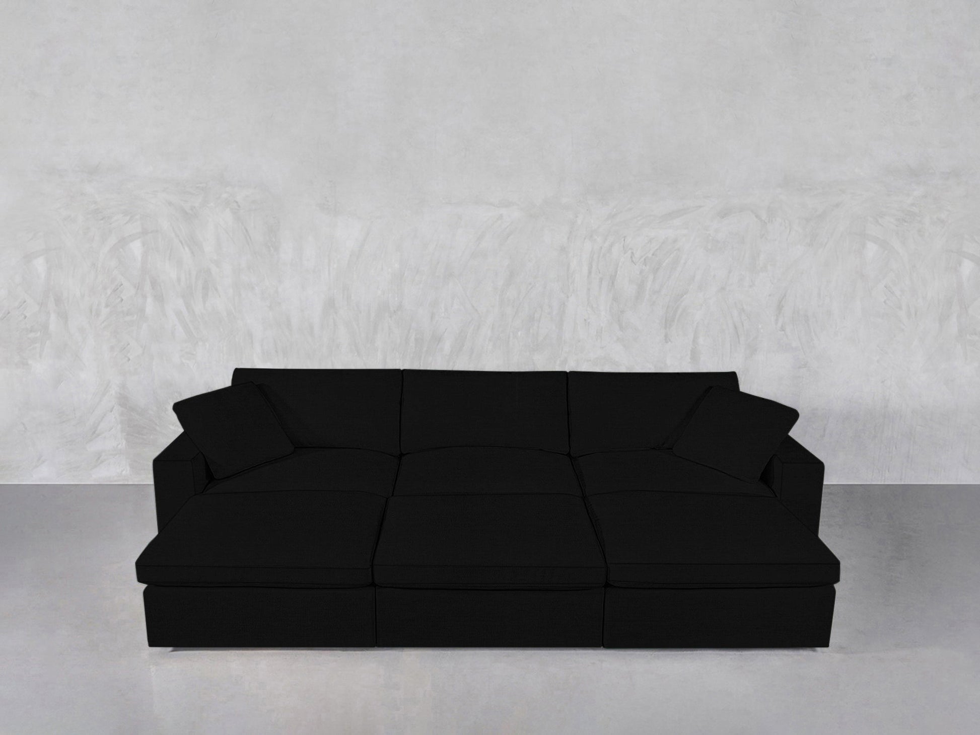 A modern black 6-seat modular daybed with two matching black throw pillows sits against a textured light gray wall on a polished gray floor.
