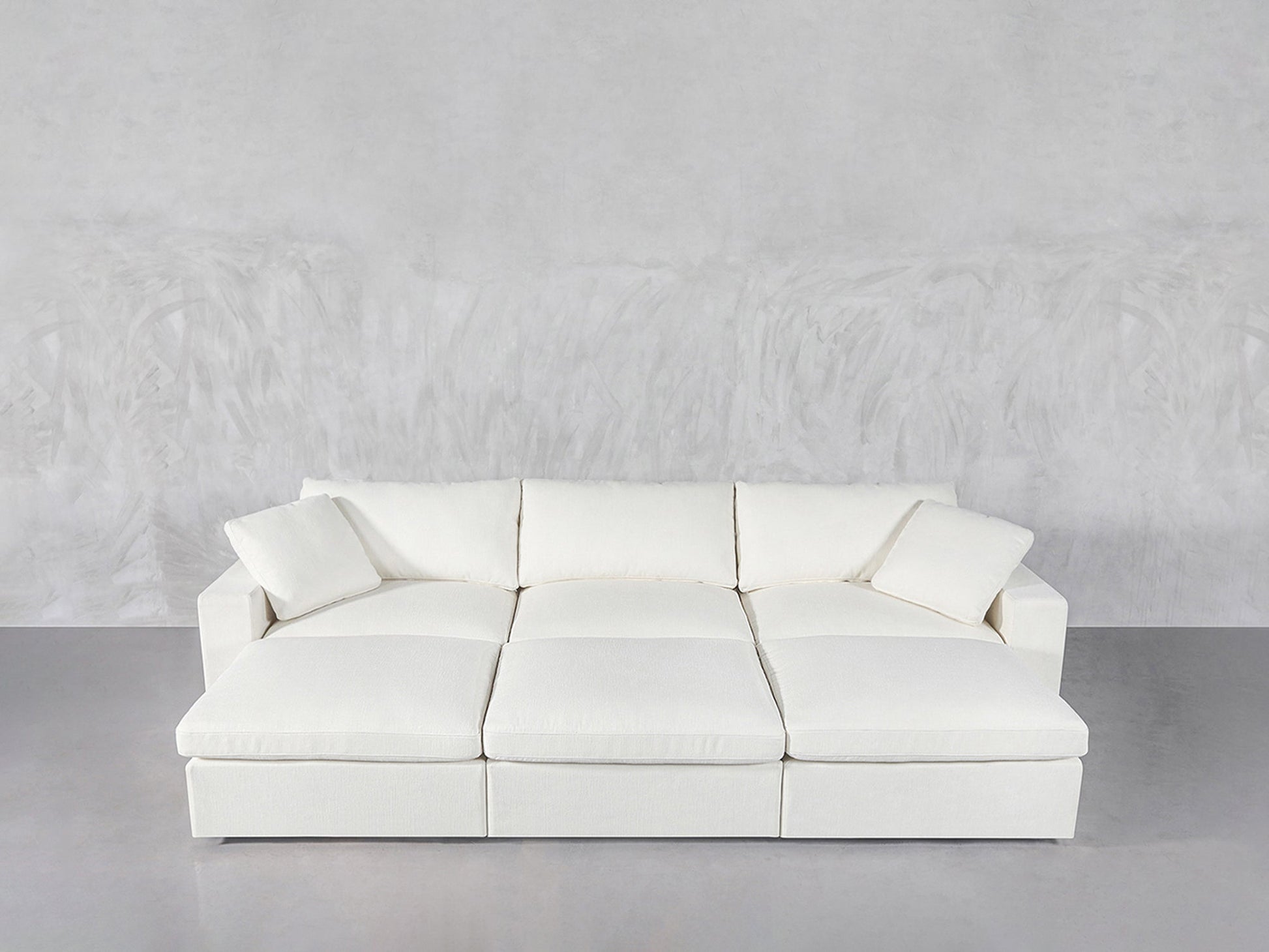 A modern, white 6-Seat Modular Daybed with wide cushions and two matching throw pillows, set against a light gray textured wall and floor.