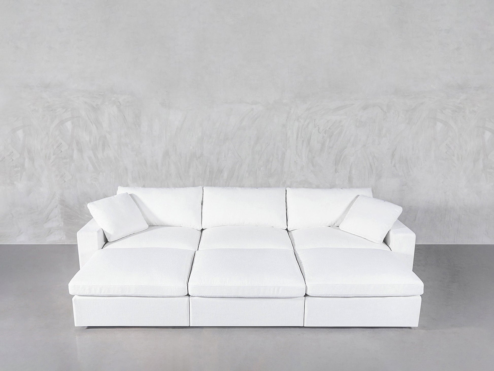 A 6-Seat Modular Daybed in modern white with three seat cushions and two matching throw pillows sits against a light gray textured wall on a smooth gray floor.