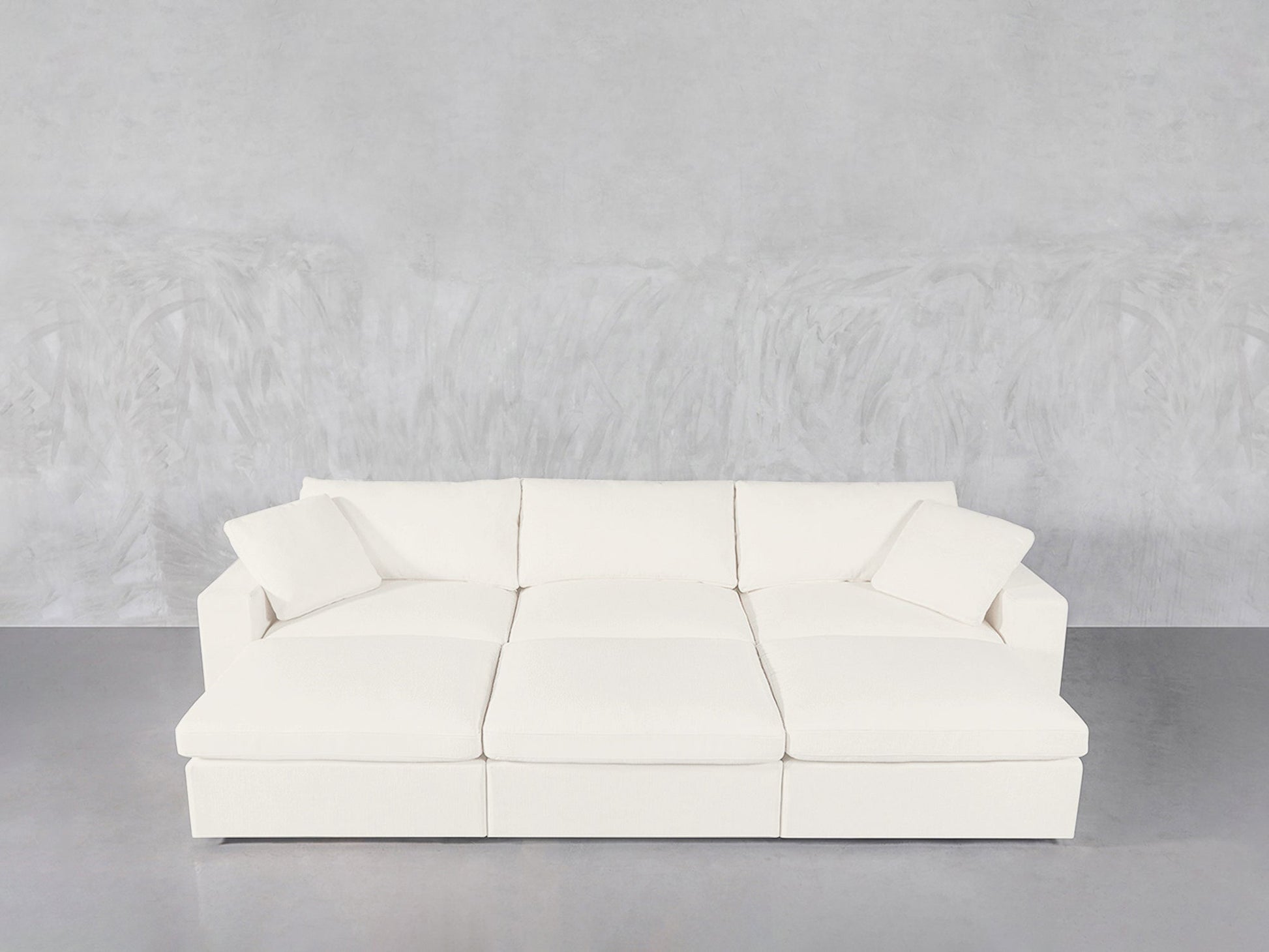 A modern 6-Seat Modular Daybed in white, featuring six seats and two matching square cushions, set against a light gray textured wall and smooth gray floor.