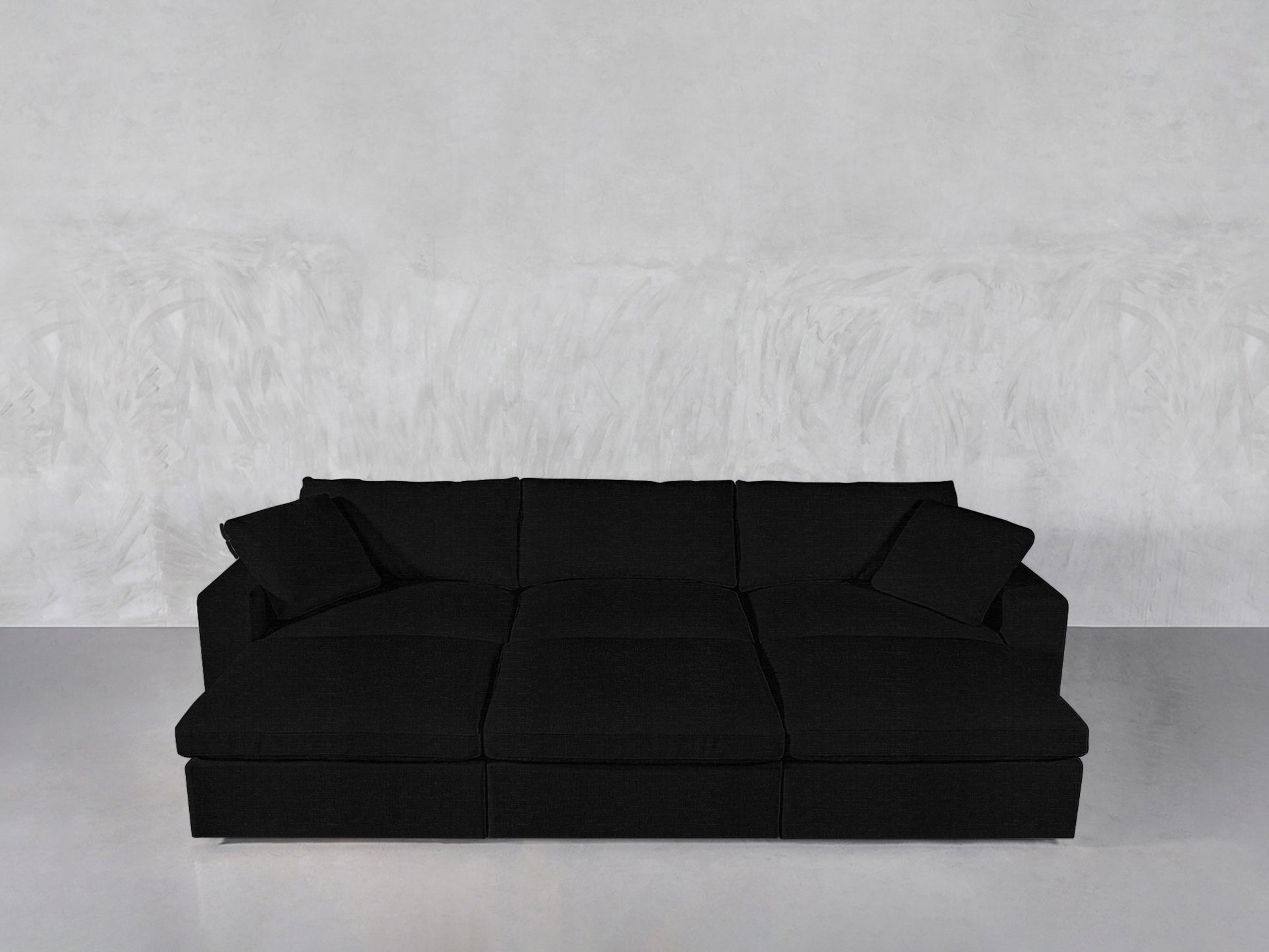 A modern black 6-Seat Modular Daybed with three seat cushions, three back cushions, and two matching throw pillows is placed against a light gray textured wall and floor.
