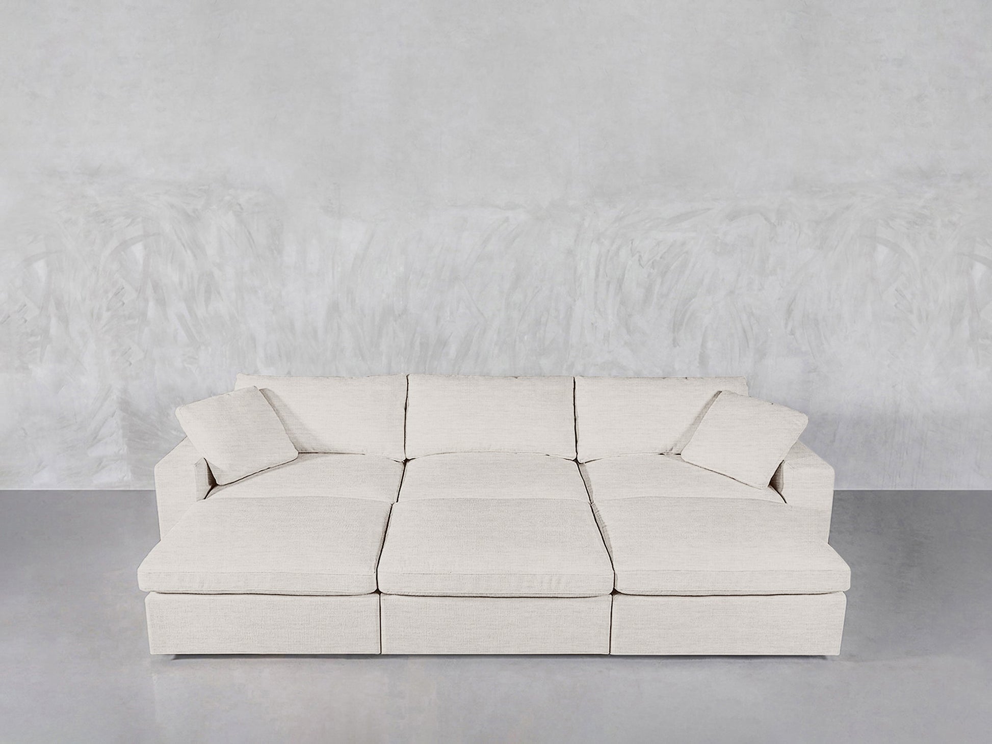 A 6-Seat Modular Daybed in light beige with wide seats and two matching cushions is placed against a textured light gray wall on a smooth gray floor.