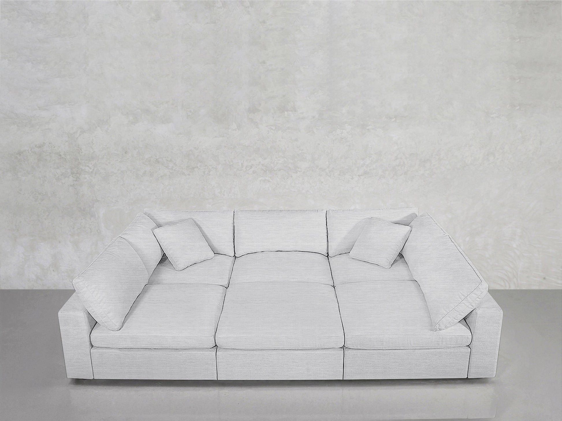 A modern 6-Seat Modular Pit in light gray, featuring three seat cushions and four matching throw pillows, is set against a plain, textured white wall on a smooth gray floor.