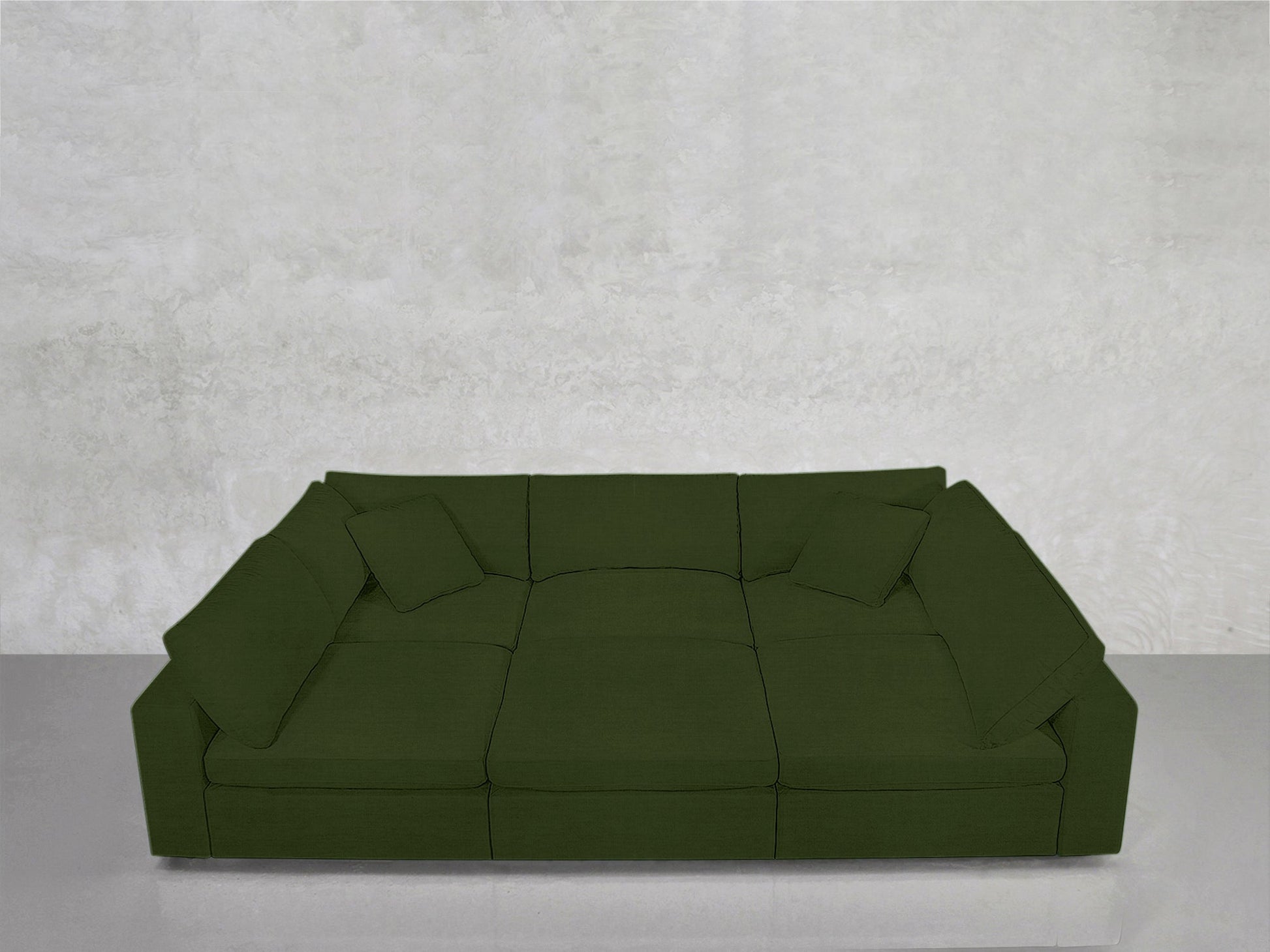 The 6-Seat Modular Pit, a large deep green sectional with three seat cushions and matching pillows, sits against a textured light gray wall on a smooth, light gray floor.