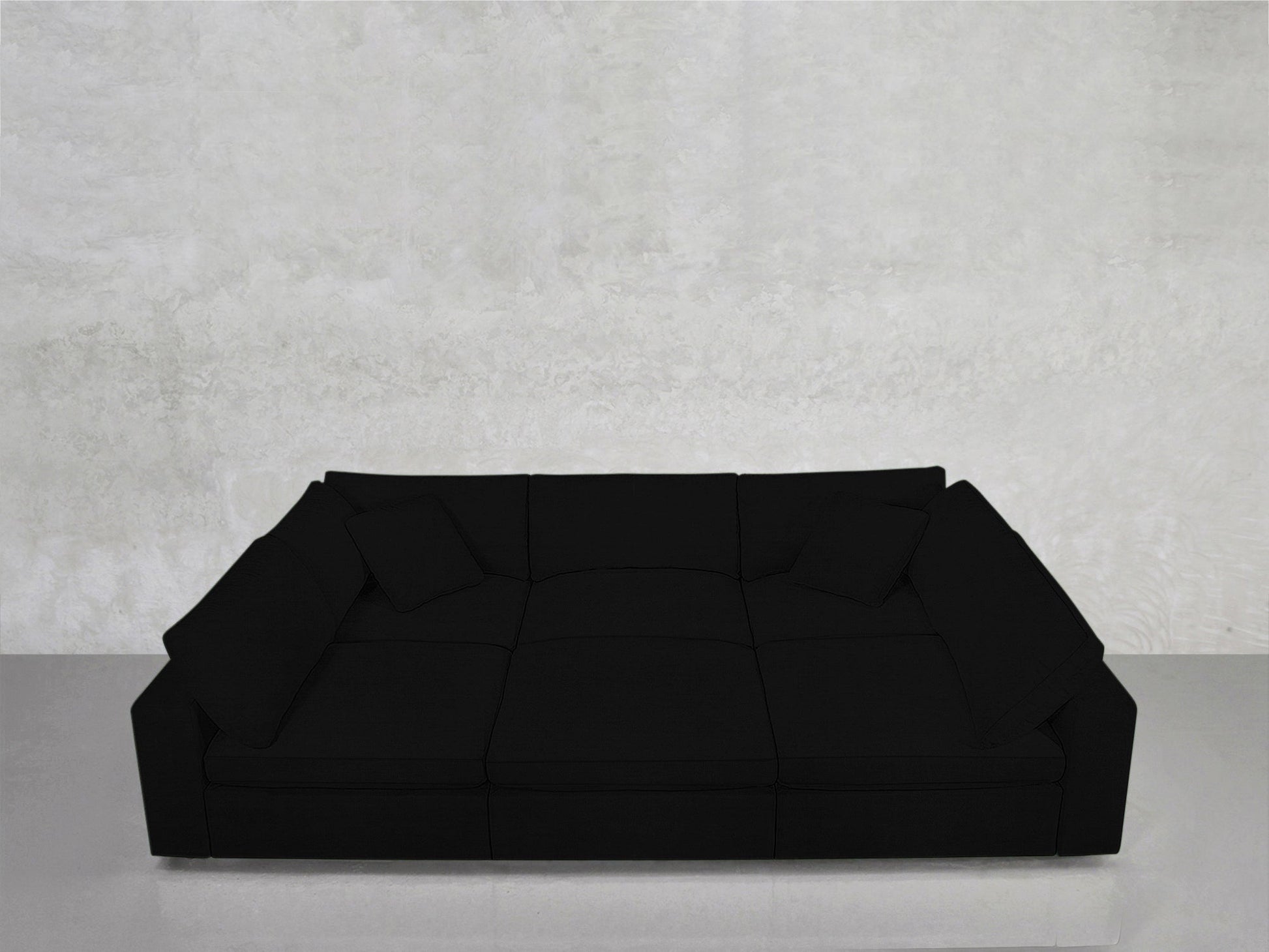 The 6-Seat Modular Pit, a spacious modern black sofa with wide armrests and four cushions, sits against a light gray textured wall on a smooth, reflective floor.