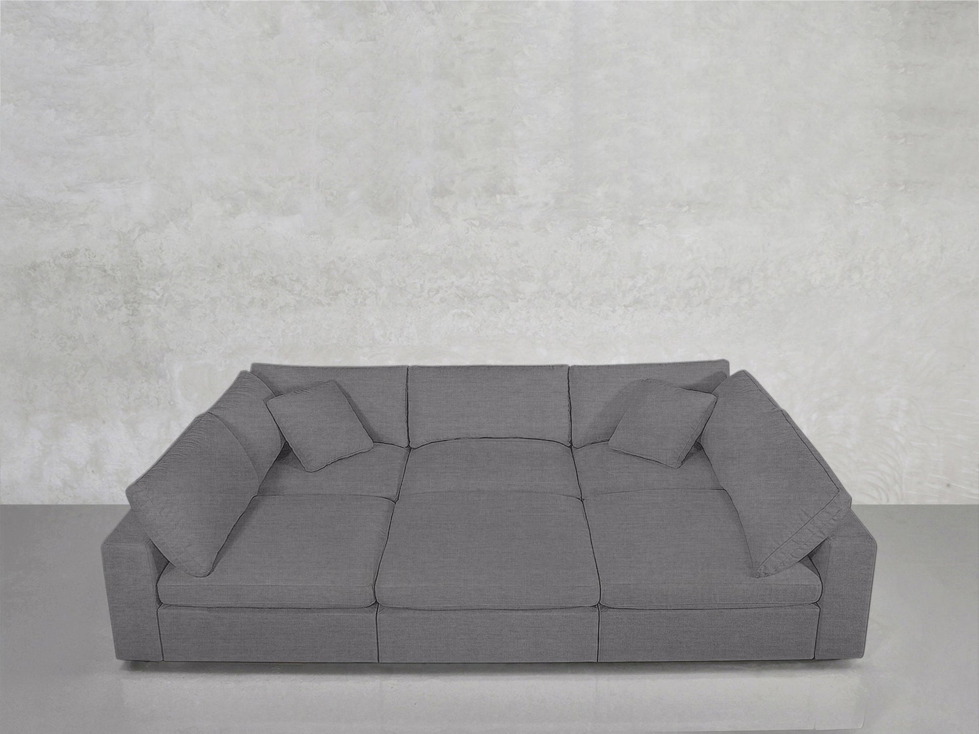 The 6-Seat Modular Pit, a spacious modern gray sofa with squared cushions and three matching throw pillows, sits against a light gray textured wall and floor.