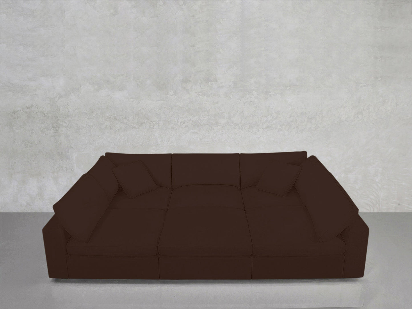 The 6-Seat Modular Pit, a spacious dark brown sectional with four matching cushions, sits against a plain light gray concrete wall atop a reflective floor.