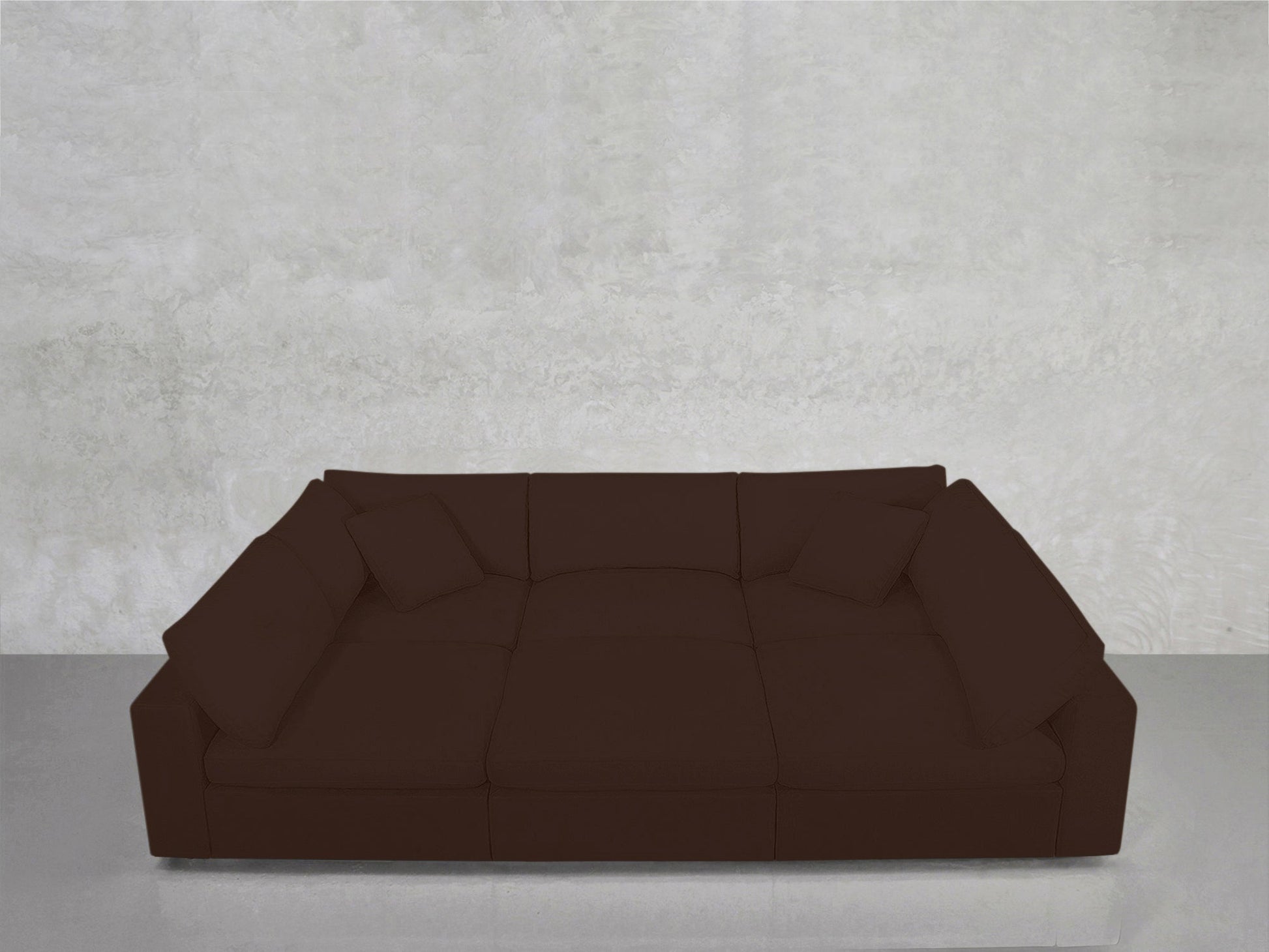 The 6-Seat Modular Pit, a spacious dark brown sectional with four matching cushions, sits against a plain light gray concrete wall atop a reflective floor.