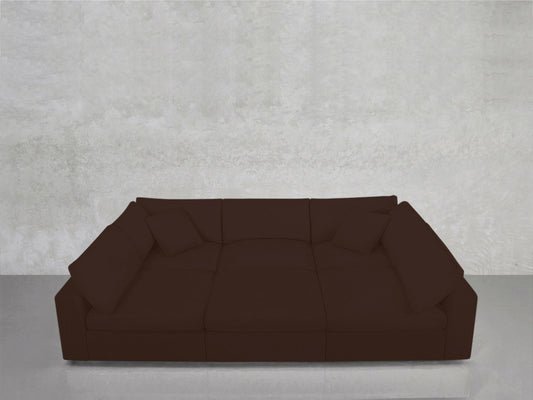 The 6-Seat Modular Pit, a spacious dark brown sectional with four matching cushions, sits against a plain light gray concrete wall atop a reflective floor.