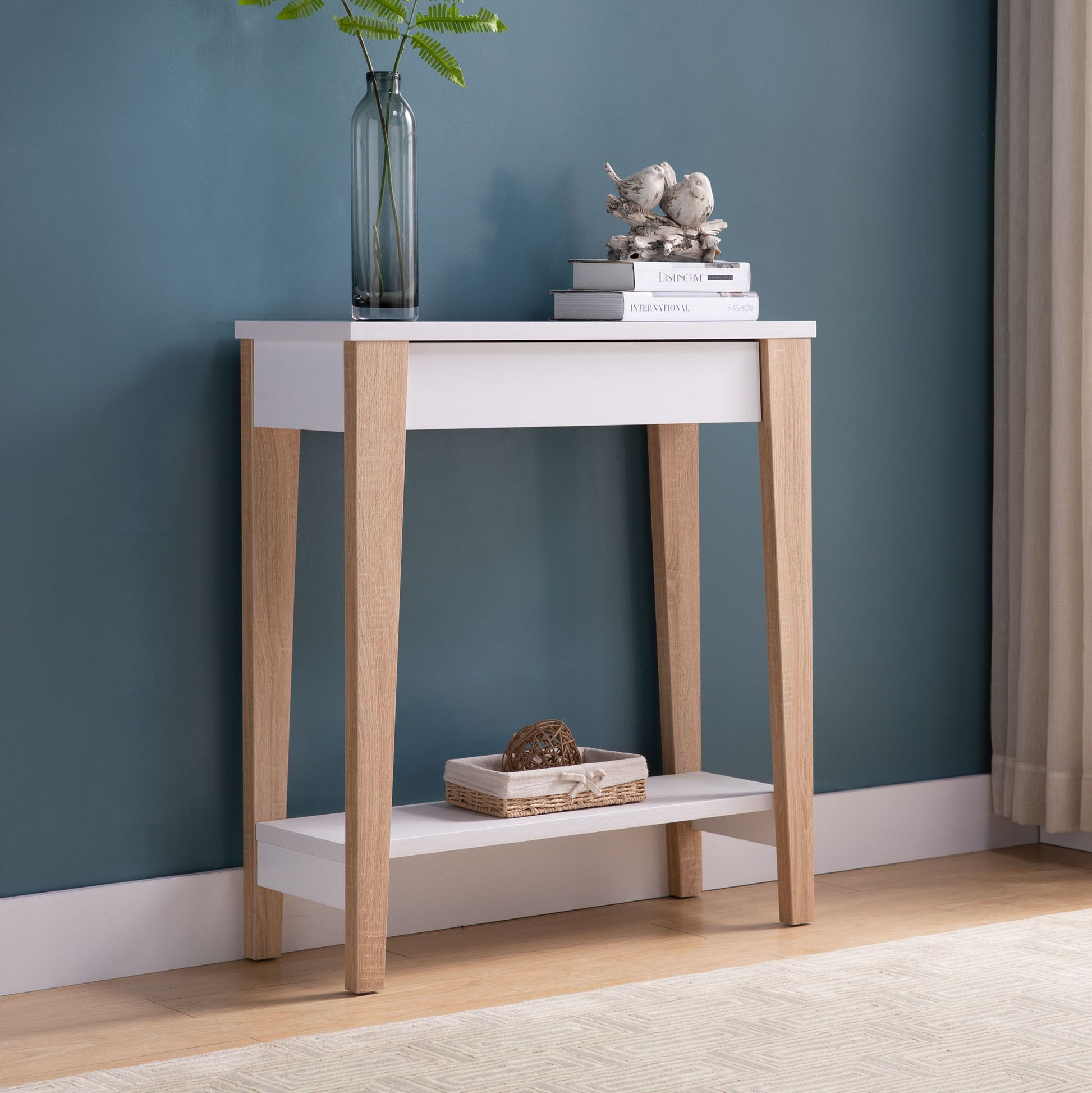The Modern Two Toned Console With Two Shelves features light wood legs and white surfaces, styled with a glass vase of green leaves, bird figurines on books, and a woven basket below, set against a blue wall.