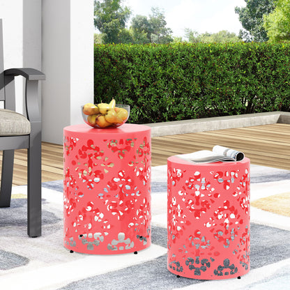 Two Outdoor Metal Modern Design End Tables in pink with cutout patterns sit on a patio by a chair—one holds fruit, the other a magazine. Bushes and greenery serve as the backdrop.