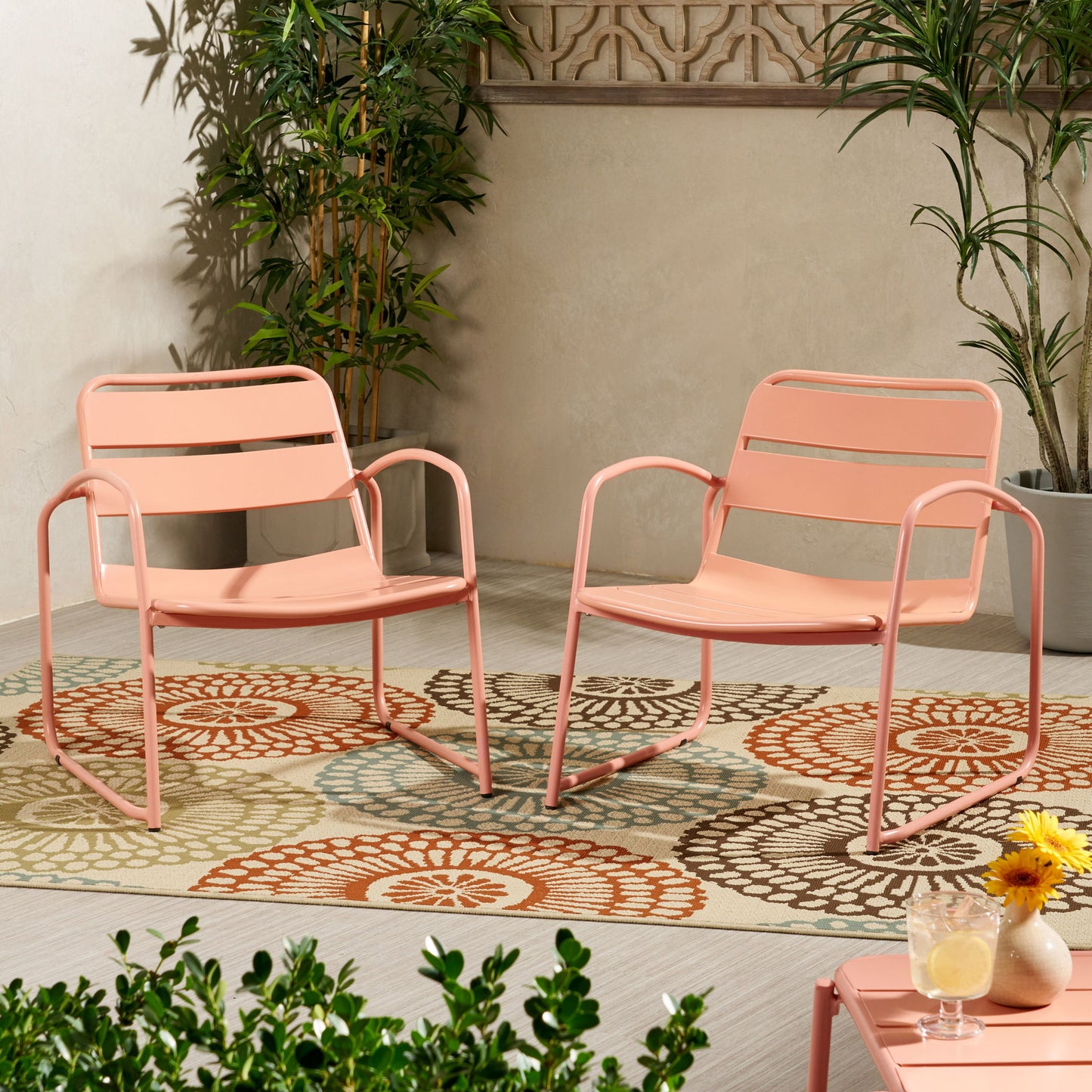 Two Conrad - Chair Outdoor Seating pieces in pink metal sit on a floral-patterned outdoor rug atop a light wooden floor, surrounded by green plants, with a small table holding drinks and a yellow flower nearby.