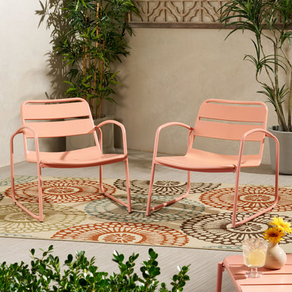 Two Conrad - Chair Outdoor Seating pieces in pink metal sit on a floral-patterned outdoor rug atop a light wooden floor, surrounded by green plants, with a small table holding drinks and a yellow flower nearby.