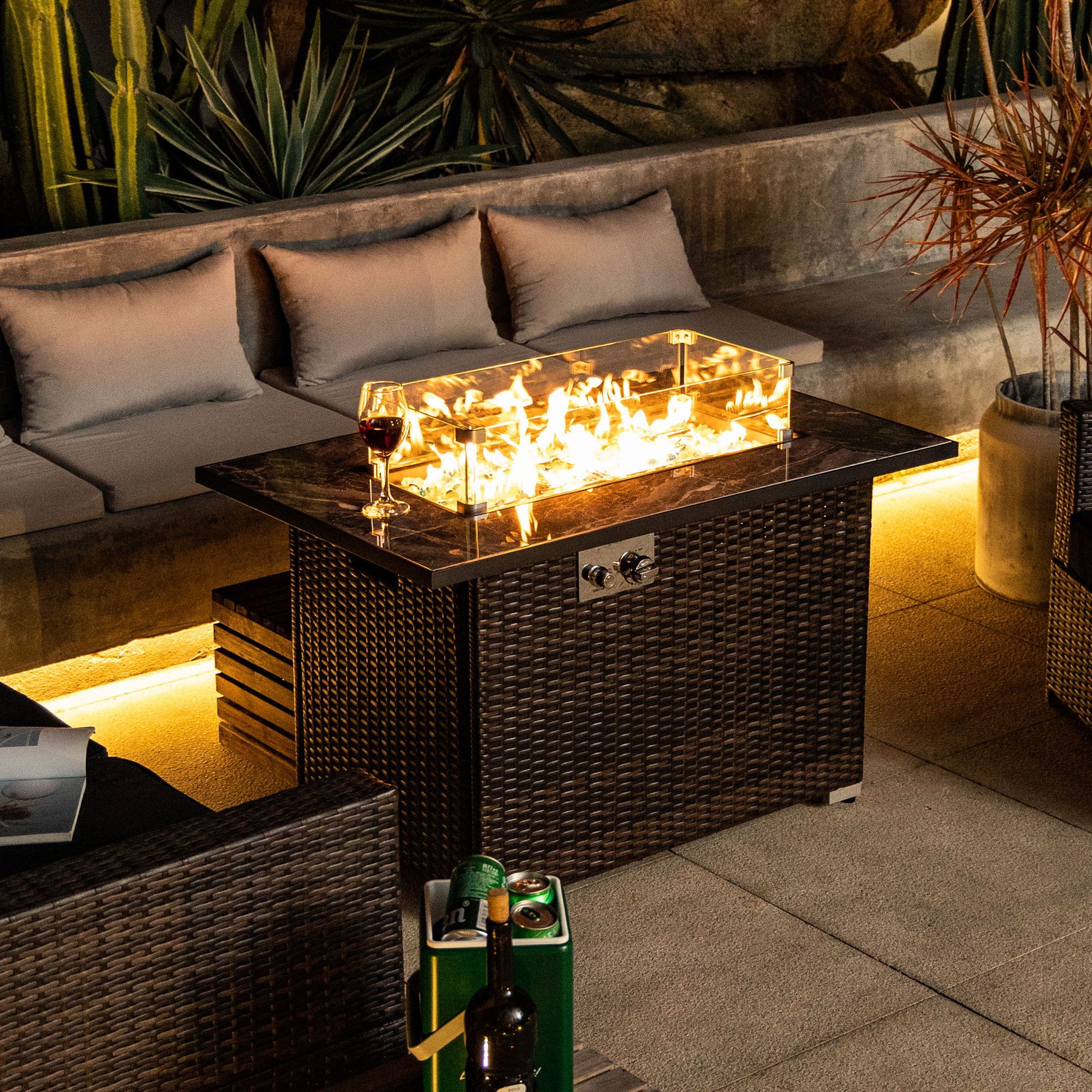 A cozy patio boasts the 44" Outdoor Fire Pit Table, Propane Fire Table with Ceramic Tabletop in Espresso. Glass sides reveal glowing flames, with a wine glass on top. Cushioned seating and lush plants complete the inviting outdoor scene.