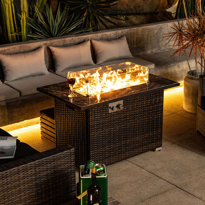 A cozy patio boasts the 44" Outdoor Fire Pit Table, Propane Fire Table with Ceramic Tabletop in Espresso. Glass sides reveal glowing flames, with a wine glass on top. Cushioned seating and lush plants complete the inviting outdoor scene.