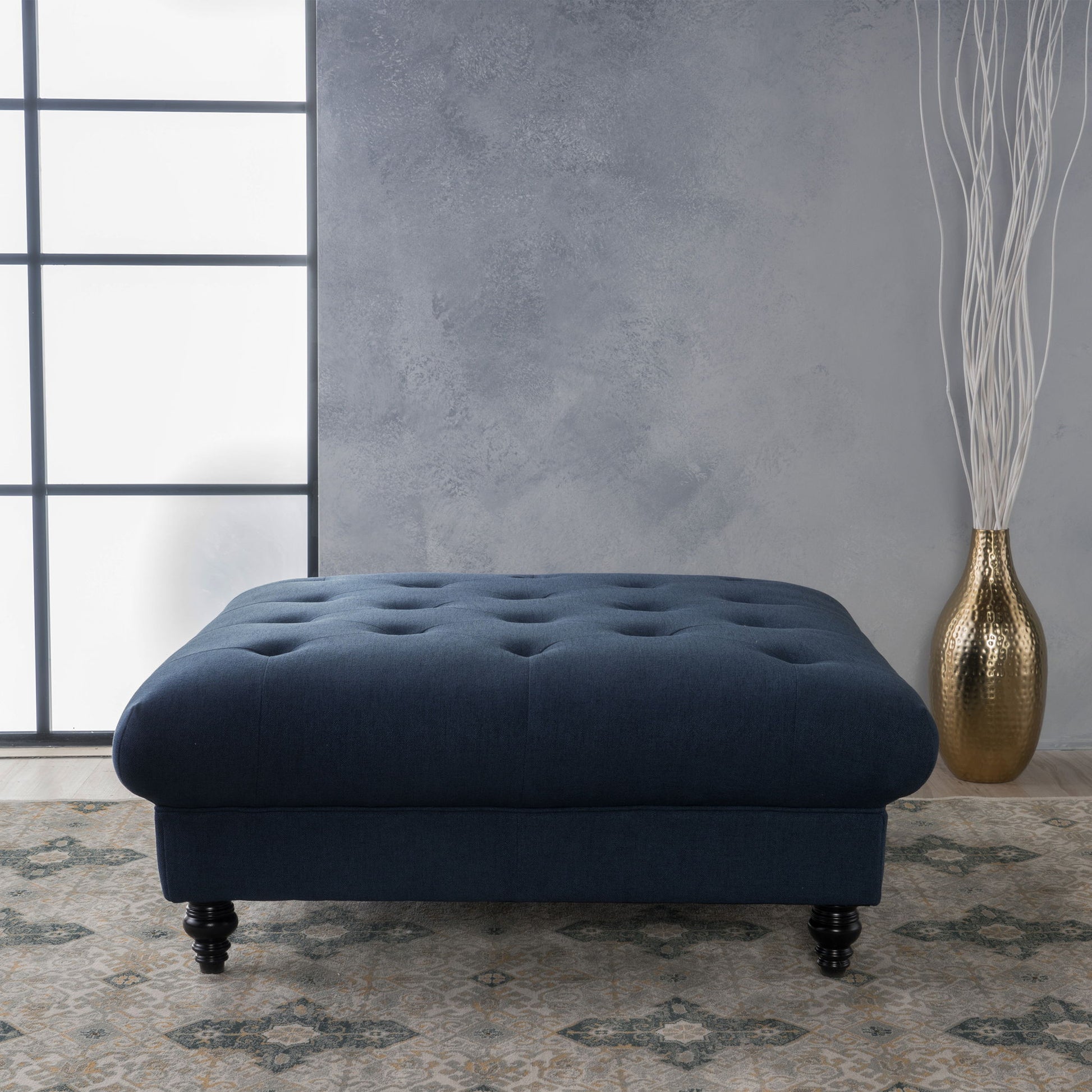 The Madrid - Tufted Ottoman Footrest in dark blue sits on a patterned rug before a gray textured wall, with a tall gold vase holding white decorative branches to its right.