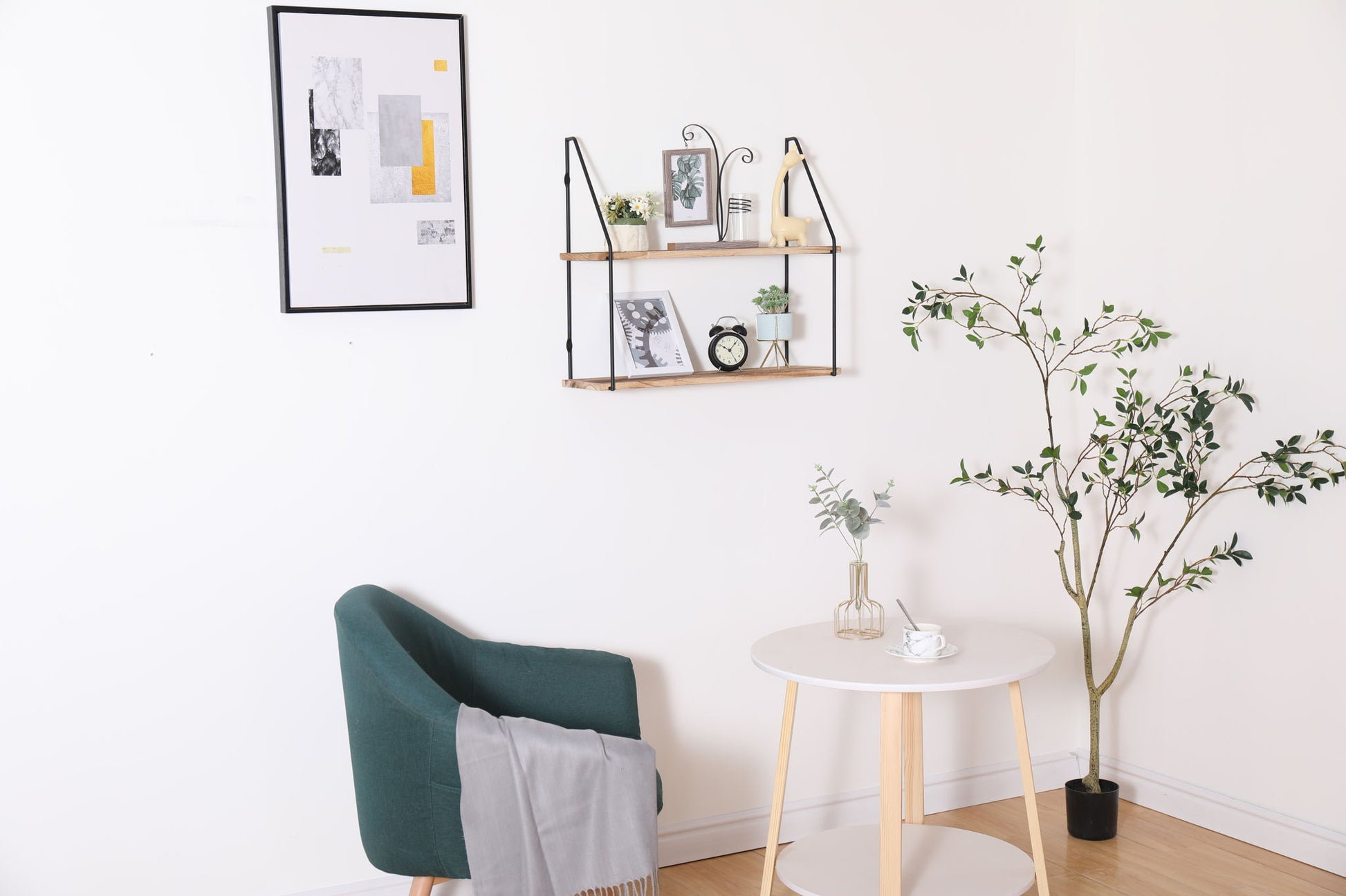 A modern, minimalistic room features a green armchair with a gray blanket, a white round table, and the Gardenia Wall Mounted Wall Storage for displaying decor, plus a framed abstract print and a potted plant in the corner.