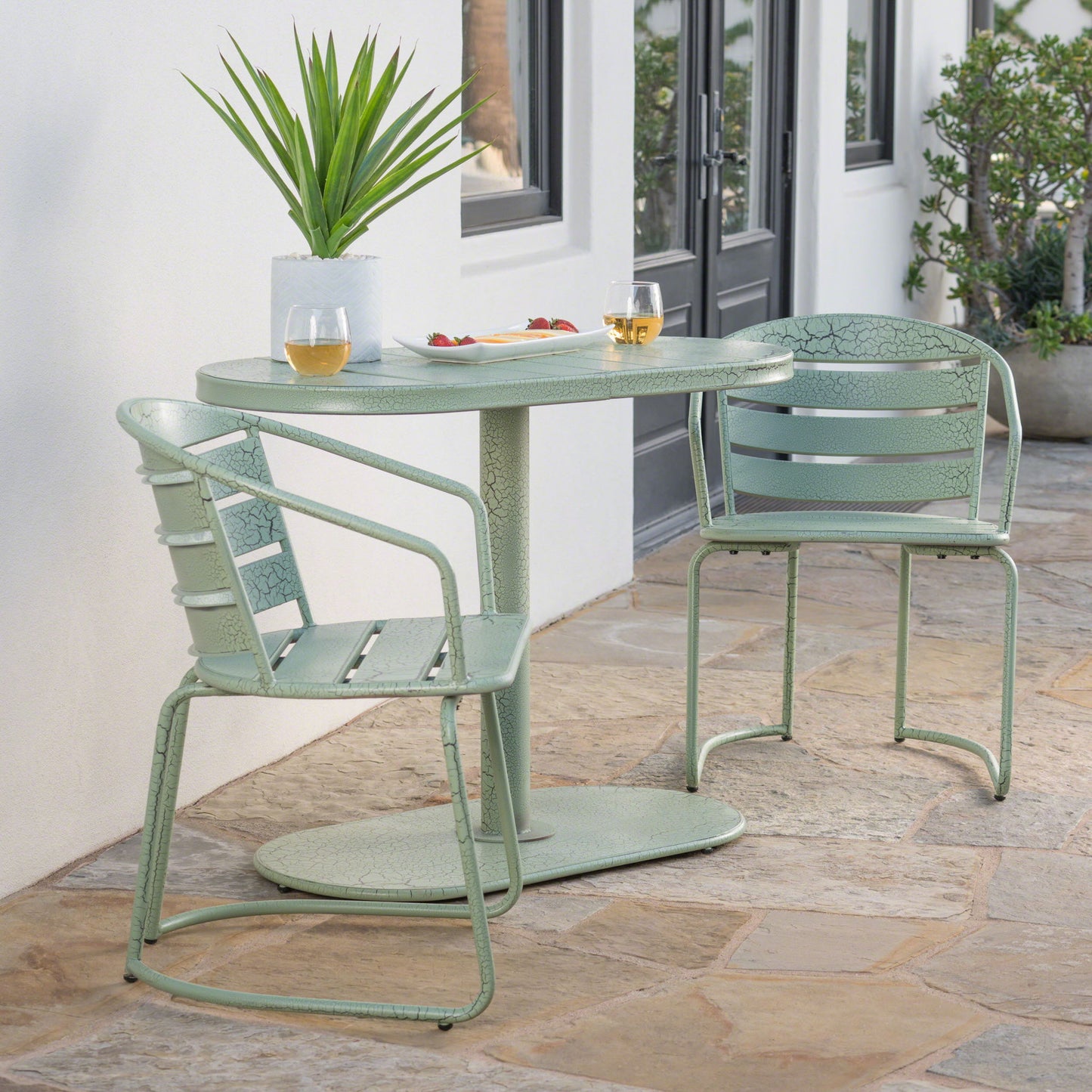 The 3 Piece Elegant Outdoor Bistro Set with 2 green metal chairs and an oval table is placed on a stone patio outside a white building, topped with two drinks, a snack plate, and a potted plant amid greenery.