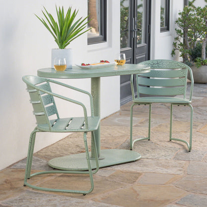 The 3 Piece Elegant Outdoor Bistro Set with 2 green metal chairs and an oval table is placed on a stone patio outside a white building, topped with two drinks, a snack plate, and a potted plant amid greenery.