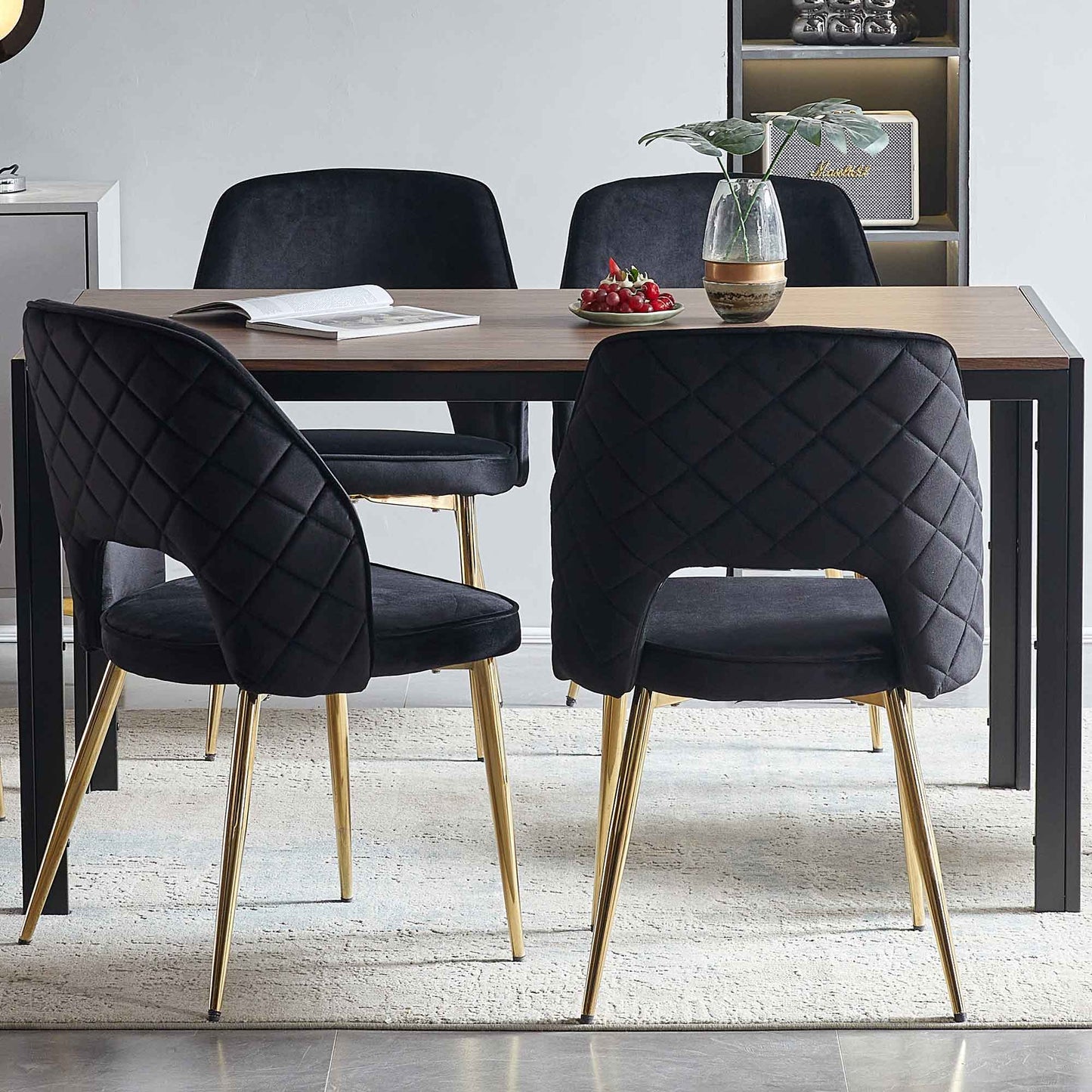 A modern dining area features a wooden table and four Velvet Dining Chairs With Metal Legs And Hollow Back, plus a small plant in a vase, an open book, and a plate of cherries on the table.